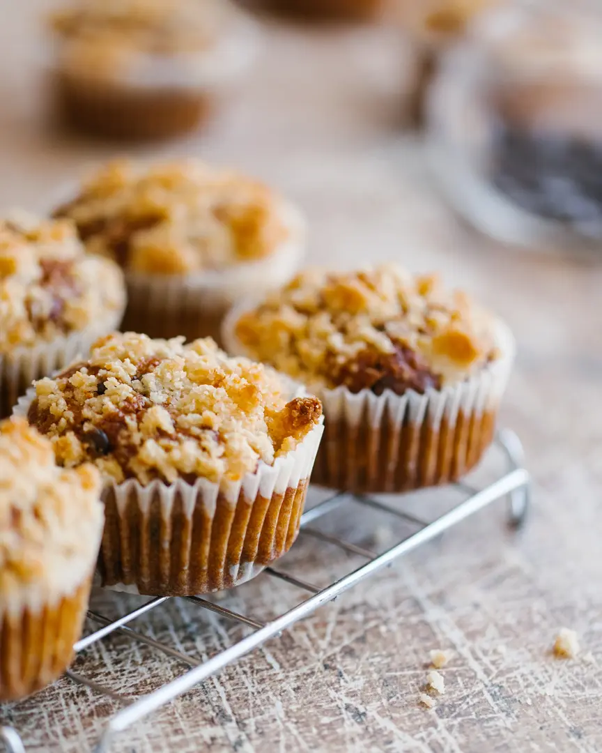 Pumpkin cupcakes. Cupcakes topped with crispy crumble are placed on a metal grate. In the background of the image, a glass jar with chocolate drops is visible in a blur.