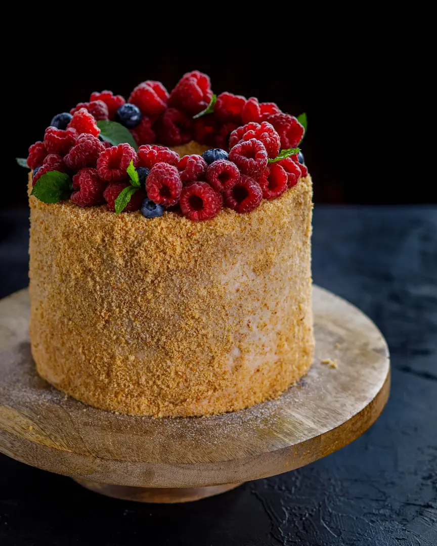 Honey cake with berries dark background. On a dark texture table, there is a brown wooden cake stand. On the cake stand is a cake sprinkled with honey crumbs. The cake is topped with a ring of berries and mint