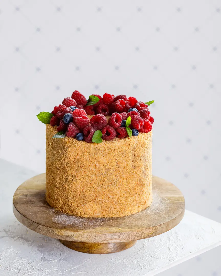 Honey cake with berries. On a white texture table, there is a brown wooden cake stand. 
On the cake stand is a cake sprinkled with honey crumbs. The cake is topped with a ring of berries and mint