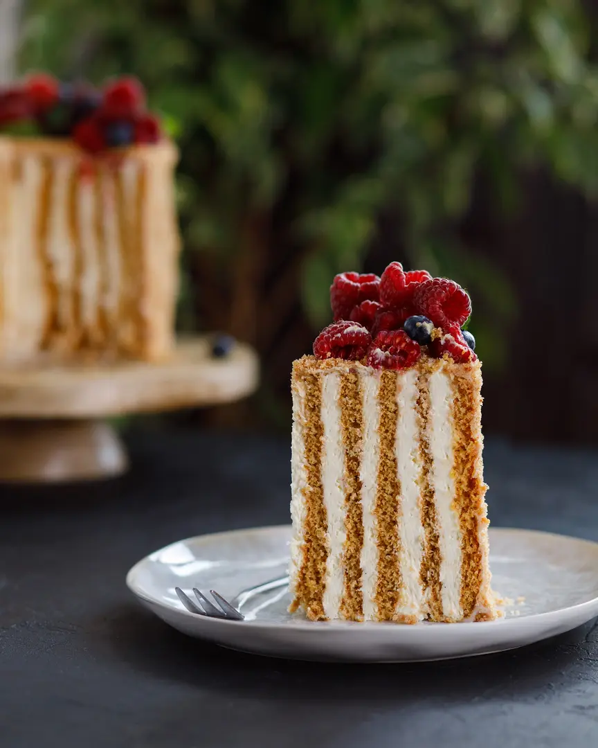 Cut of cake. On a dark texture table, there is a light plate. On the plate is a cut of cake sprinkled with honey crumbs. The cake is topped with berries and mint.
The attention of the viewer is provided with a non-standard - vertical arrangement of cake layers in the cake.