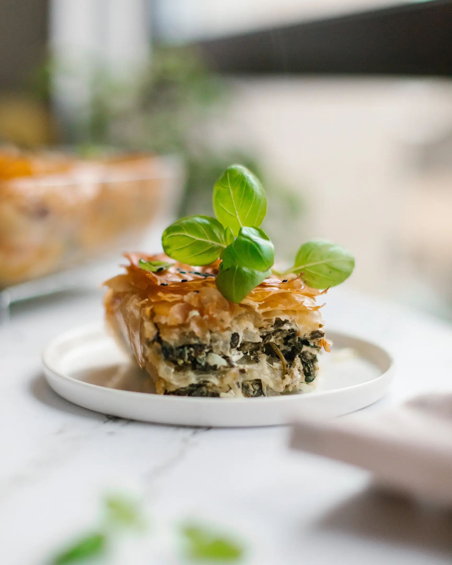 A piece of Greek spanakopita pie is on a plate. A piece of Greek spanakopita pie is on a plate, with a basil leaf on top.
Greens and the main part of the pie are visible in the background.