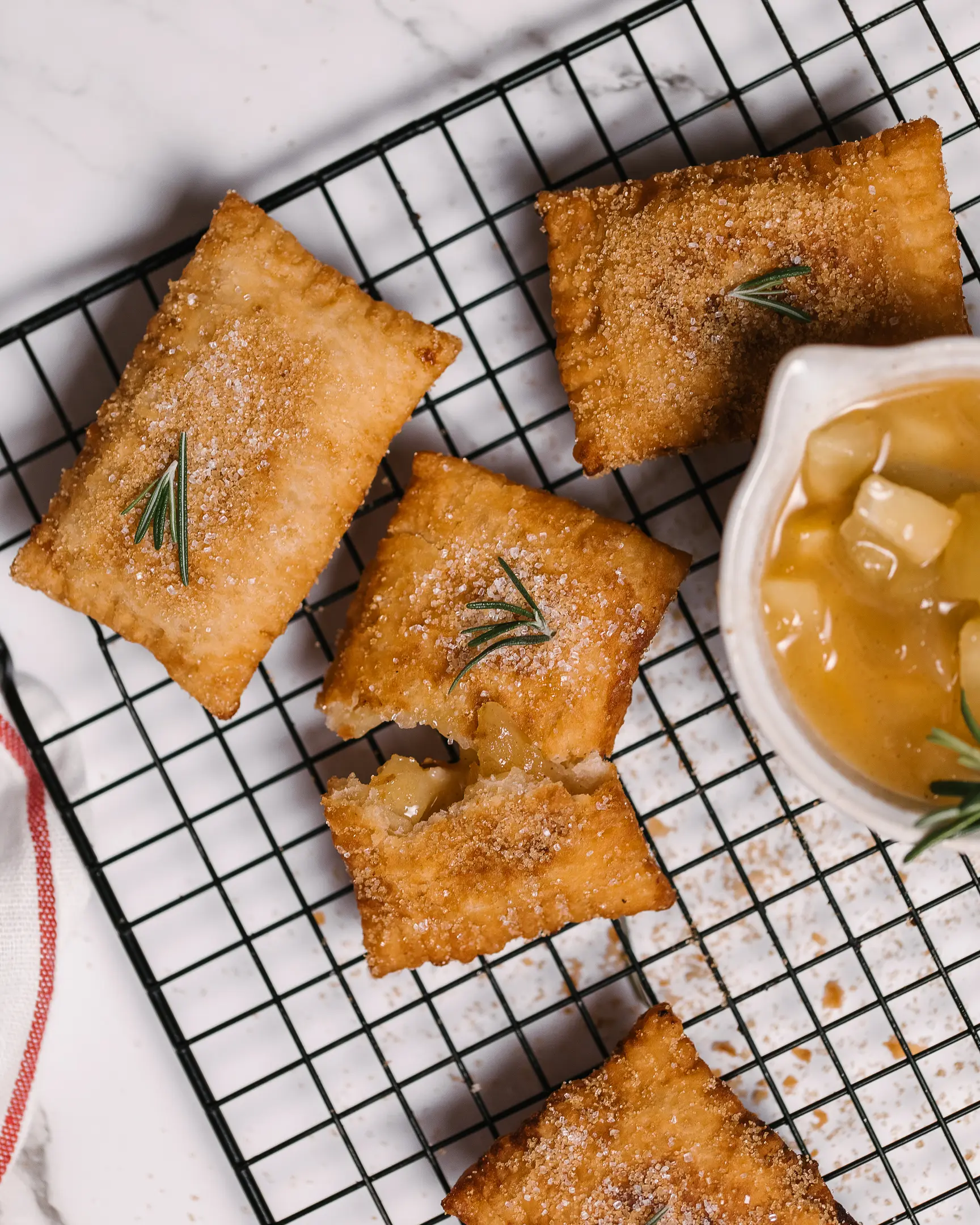 There are hand pies with apples on the pastry rack.  There are hand pies with apples on the pastry rack. They are decorated with rosemary and cinnamon sugar. In the right corner of the photo, there is a plate filled with apples.