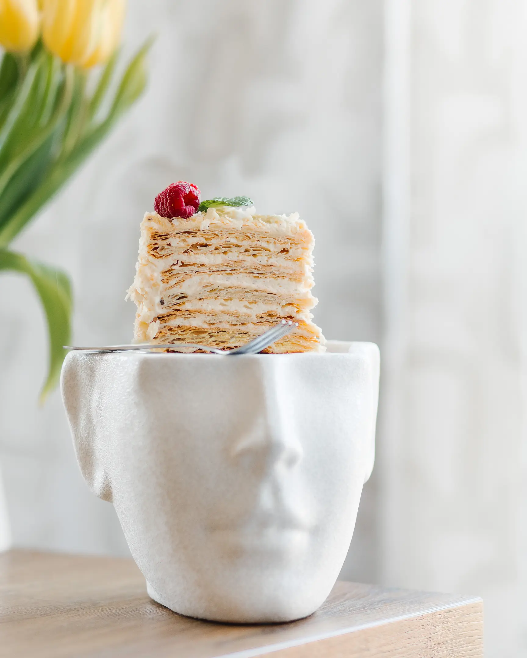 A piece of cake Napoleon. A piece of puff cake Napoleon stands on a plate in the form of a human head. The cake is on the table. In the background are flowers in a jar