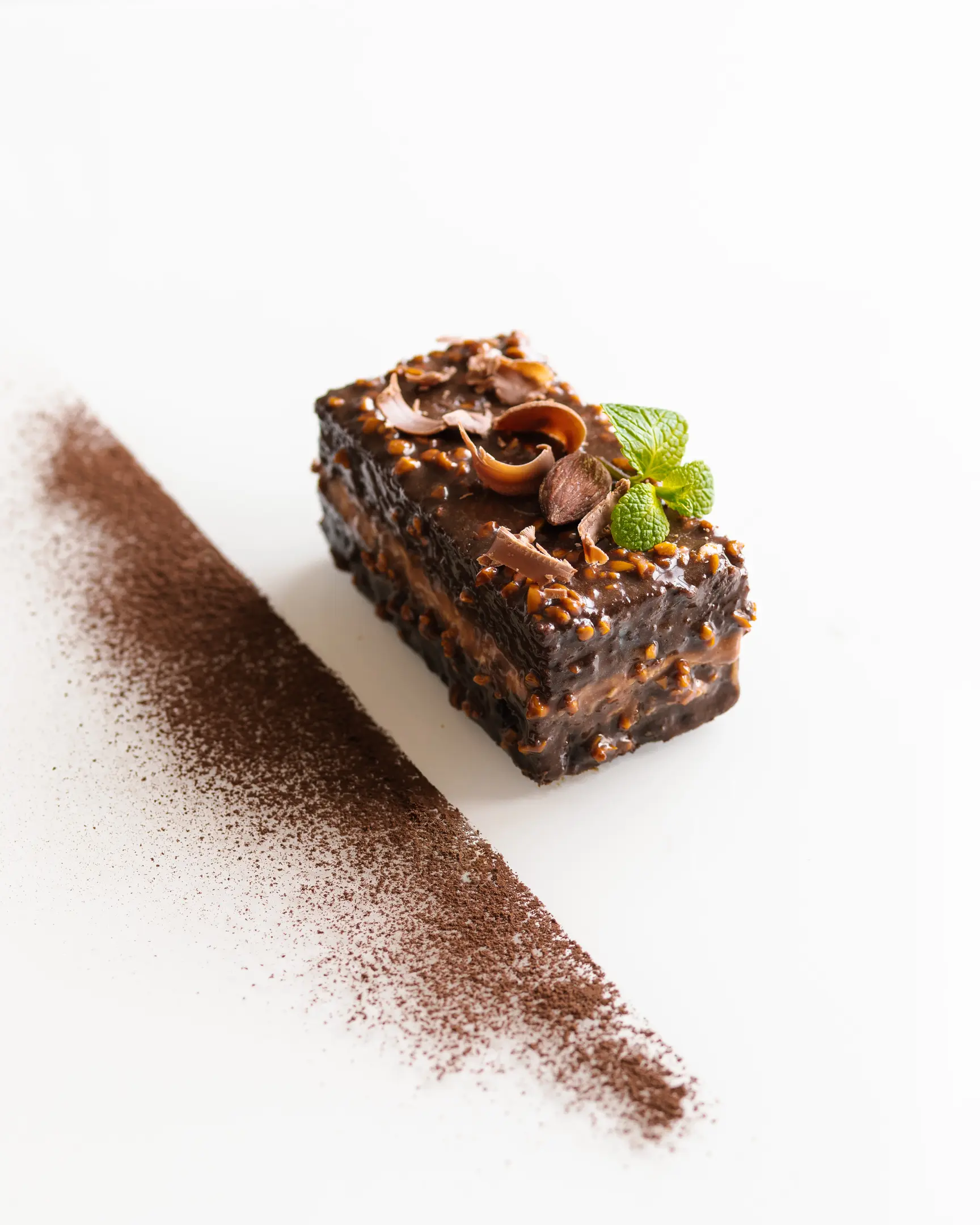 On a light glass surface lies a chocolate cake decorated with cacao On a light glass surface lies a chocolate cake decorated with cacao. To the left of it is a strip of cocoa. The dessert is decorated with almonds and mint.