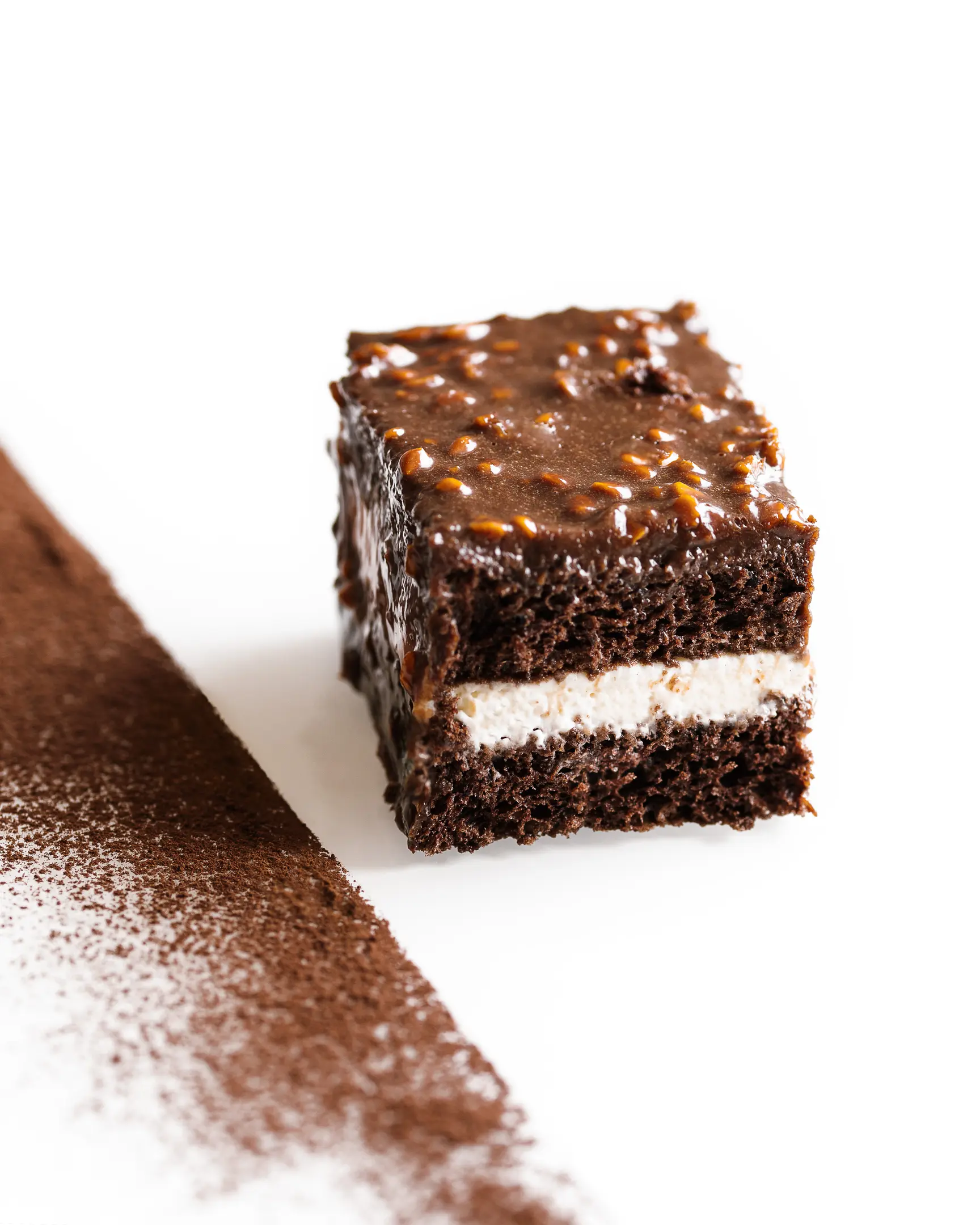 Cut of a individual cake bar. On a light glass surface lies a chocolate cake decorated with cacao. The cake is cut. To the left of it is a strip of cocoa. The dessert is decorated with almonds and mint.