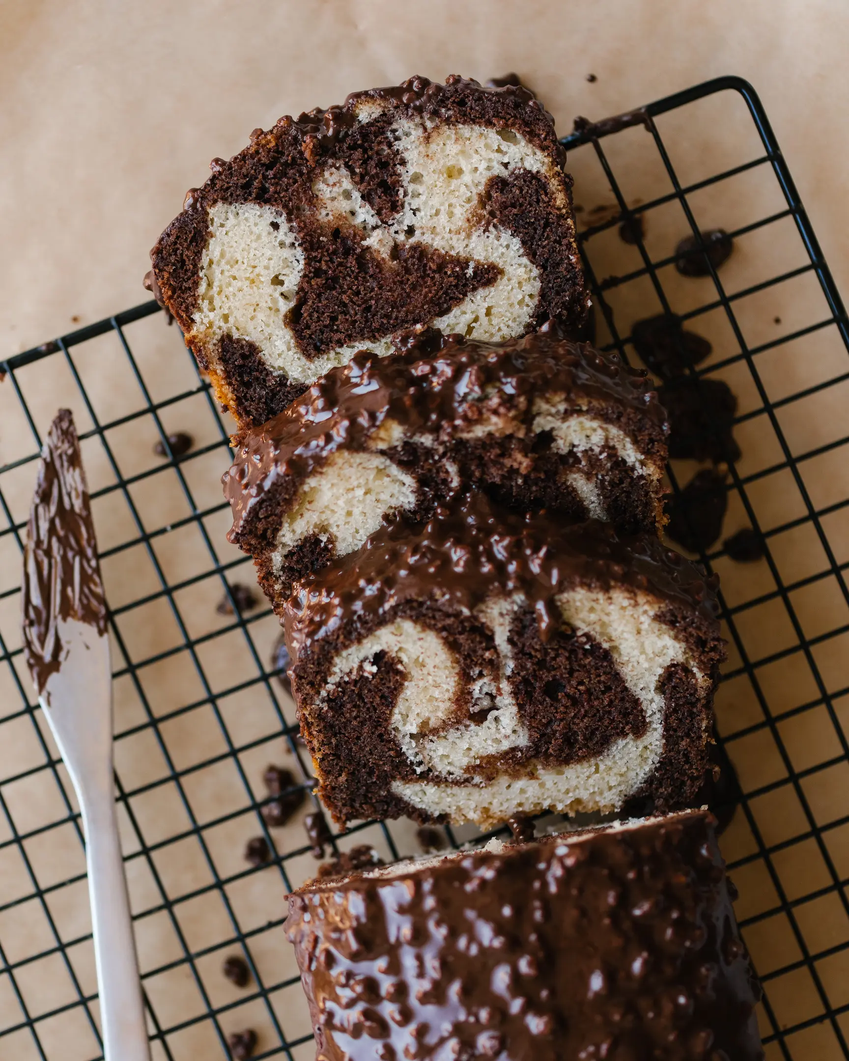 A sliced marble cake lies on a pastry rack to cool.  A sliced marble cake lies on a pastry rack to cool. The cake is cut into slices. There is a knife next to the cupcake. It's covered in chocolate icing. Drops of chocolate under the cupcake.