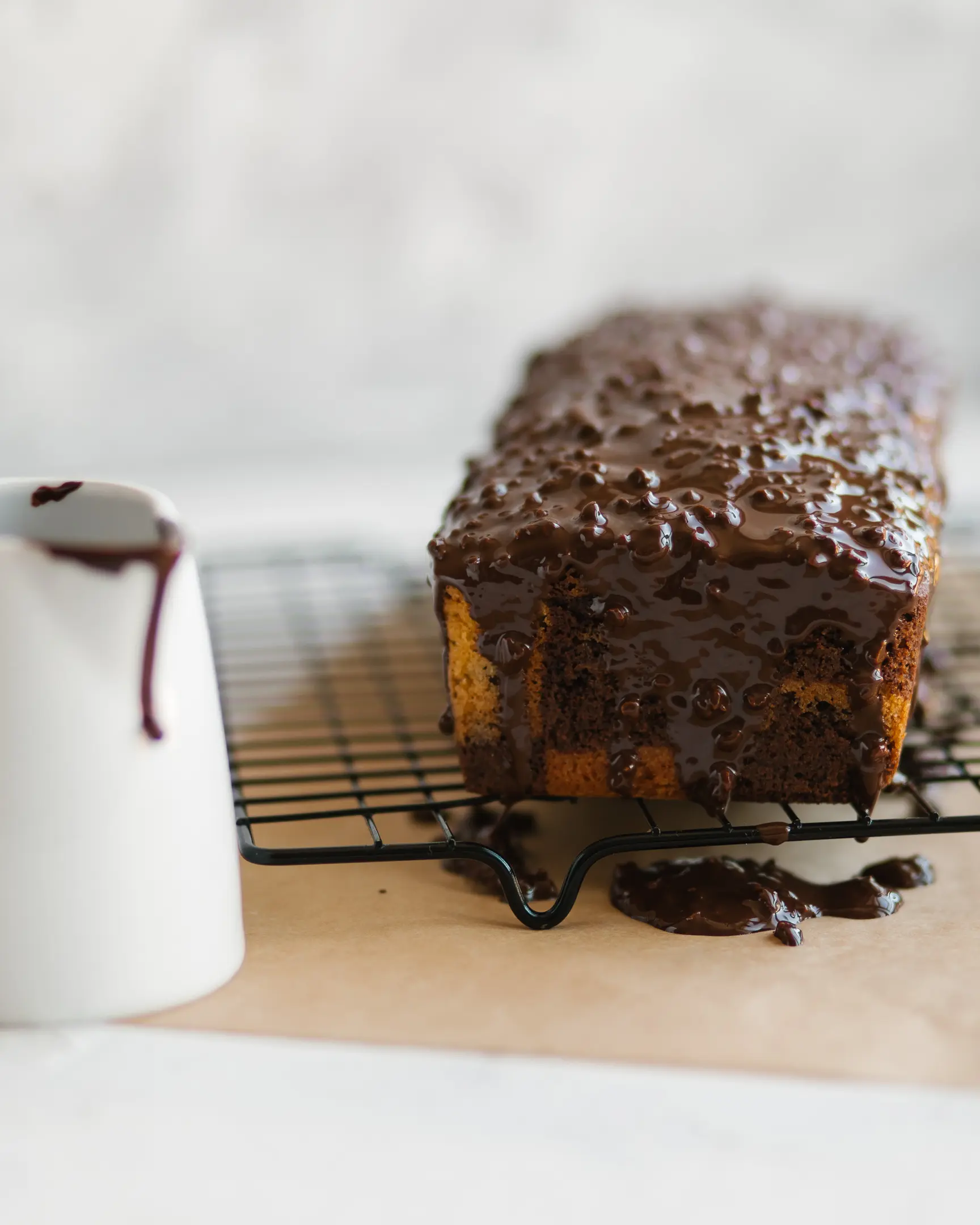 On the cake tack is a marble cupcake. I There is a pastry rack on the table. On it is a marble cupcake. It is covered with chocolate icing. Next to the cupcake is a milk jug with drops of icing dripping from it.