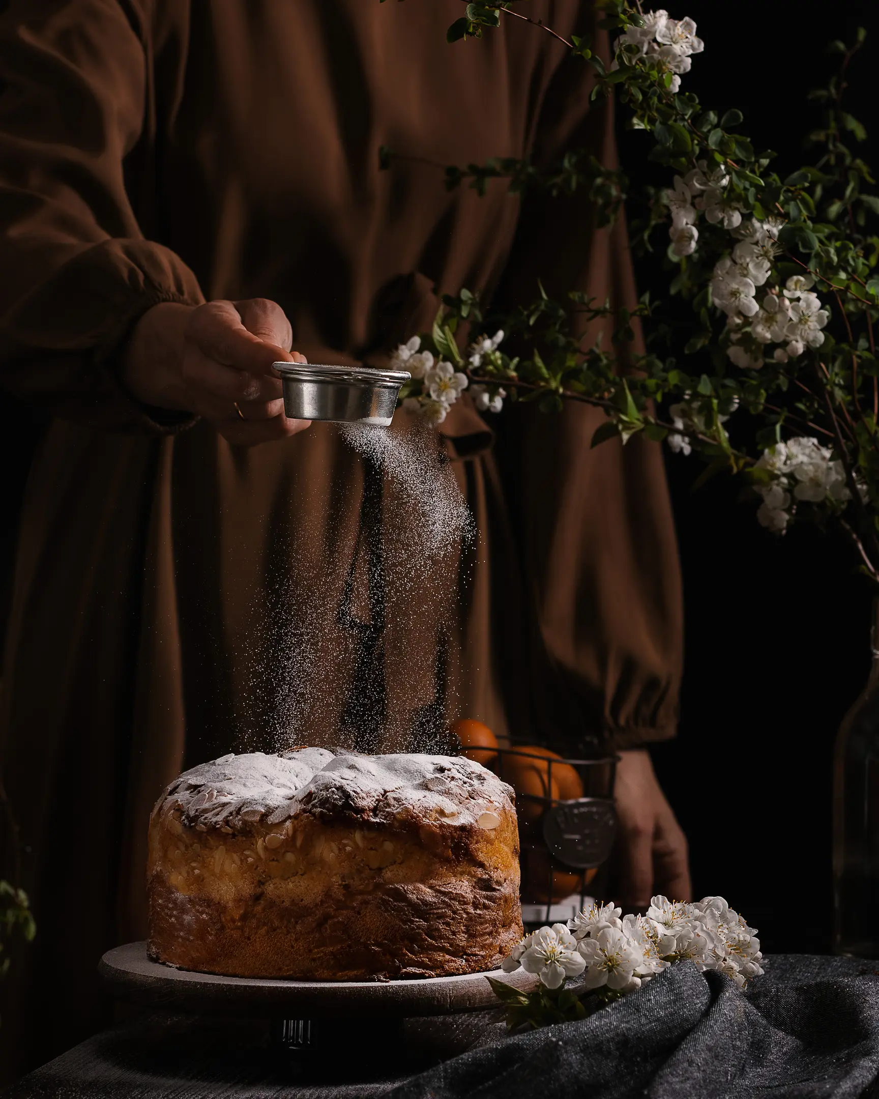 A woman sprinkles Easter bread with powdered sugar. A woman sprinkles Easter bread with powdered sugar. The bread is on the table. The table is covered with a light festive tablecloth. There are spring flowers on the table.