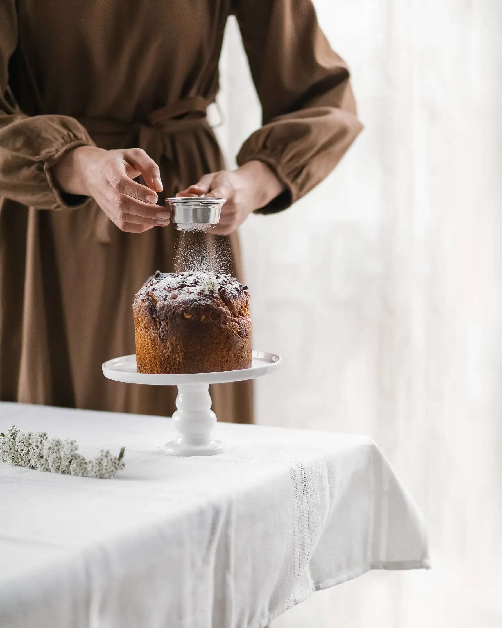 A woman sprinkles Easter bread with powdered sugar.  A woman sprinkles Easter bread with powdered sugar. The bread is on the table. The table is covered with a light festive tablecloth. There are spring flowers on the table.