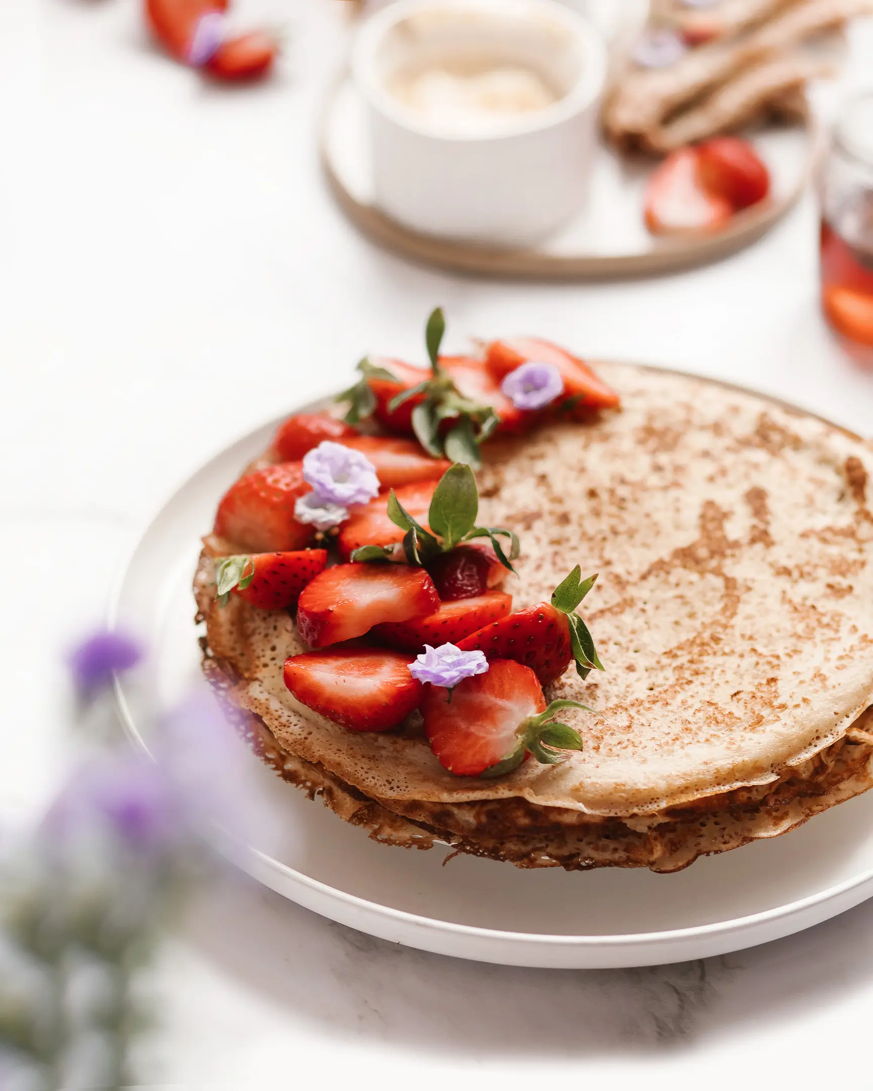 On a light background there is a plate with pancakes. On a light background, there is a plate with pancakes. in the foreground, the viewer's eye sees purple flowers. A plate of oil is visible at the back. There are strawberries on pancakes.
