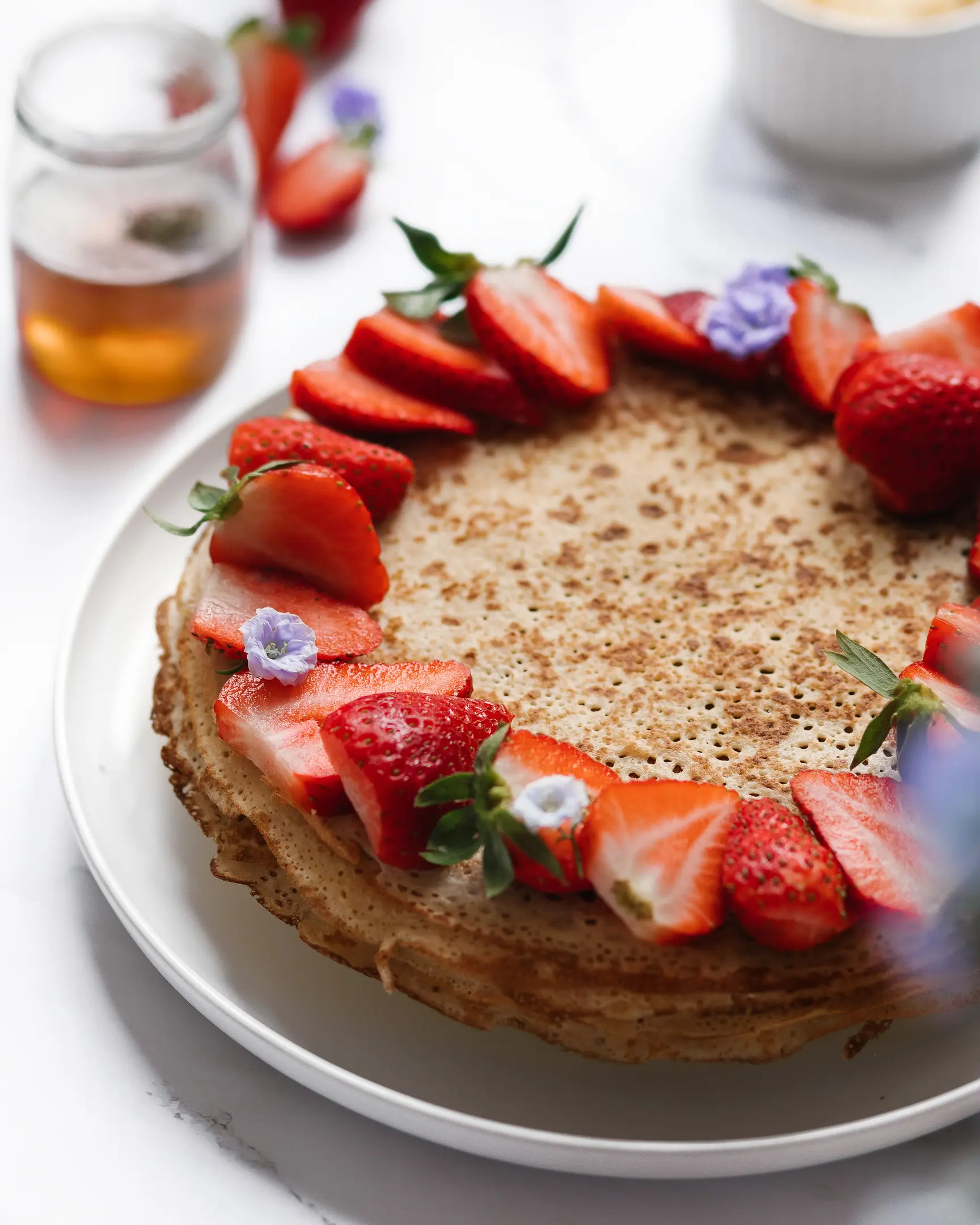 On a light background there is a plate with pancakes decorated with berries.  On a light background there is a plate with pancakes decorated with berries. in the foreground, the viewer's eye sees purple flowers. Behind you can see a plate of butter and a jar of honey. There are strawberries on pancakes.