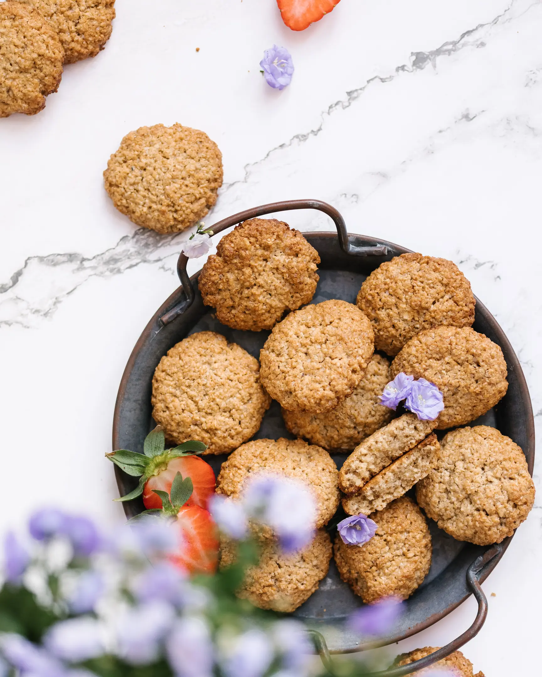 There are oatmeal cookies on a metal plate.  There are oatmeal cookies on a metal plate. In the foreground in the frame are purple flowers. Some cookies are just on the table. One of the cookies is broken.