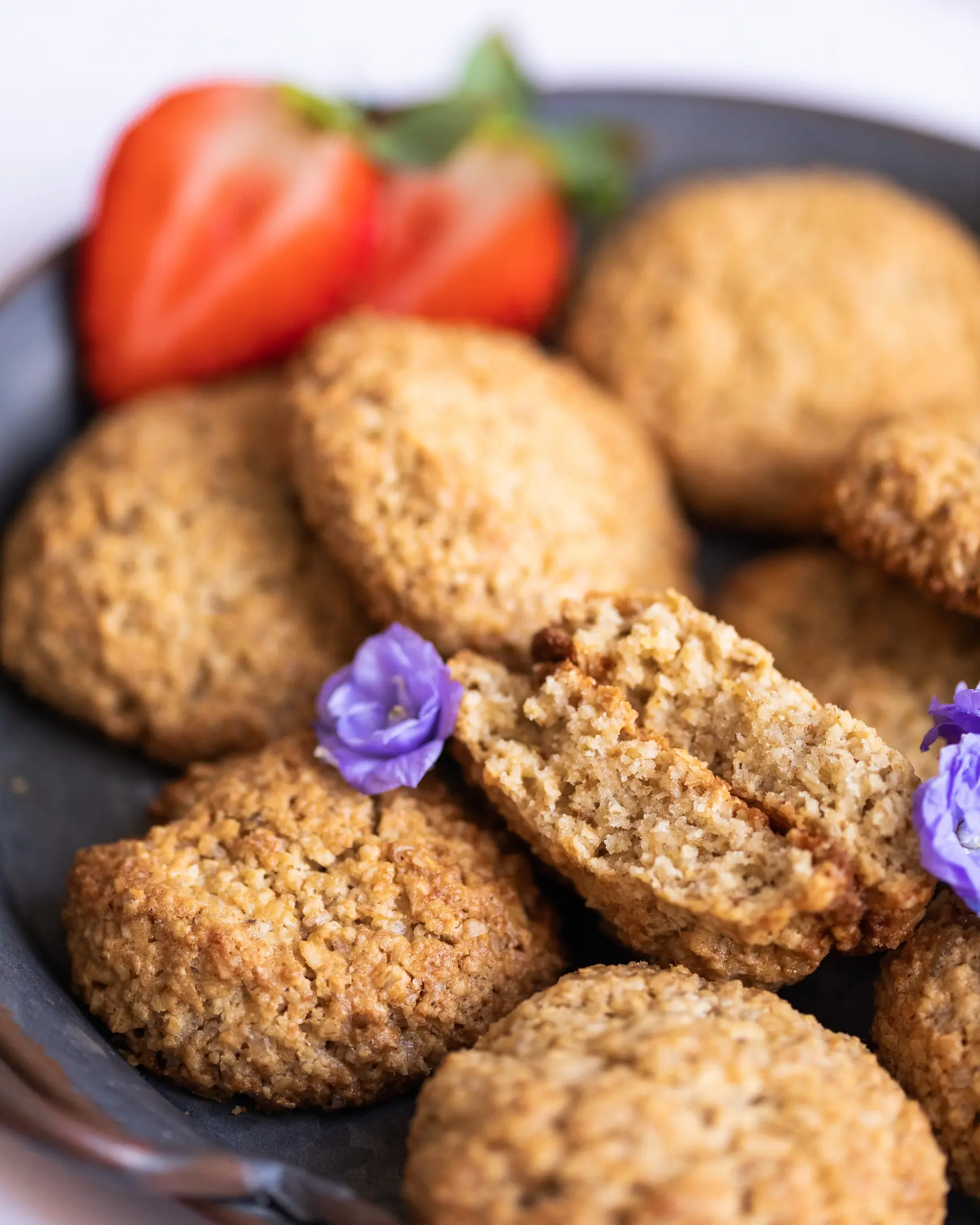 Oatmeal flourless cookies cut There are oatmeal cookies on a metal plate. In the foreground in the frame are purple flowers. Some cookies are just on the table. One of the cookies is broken.