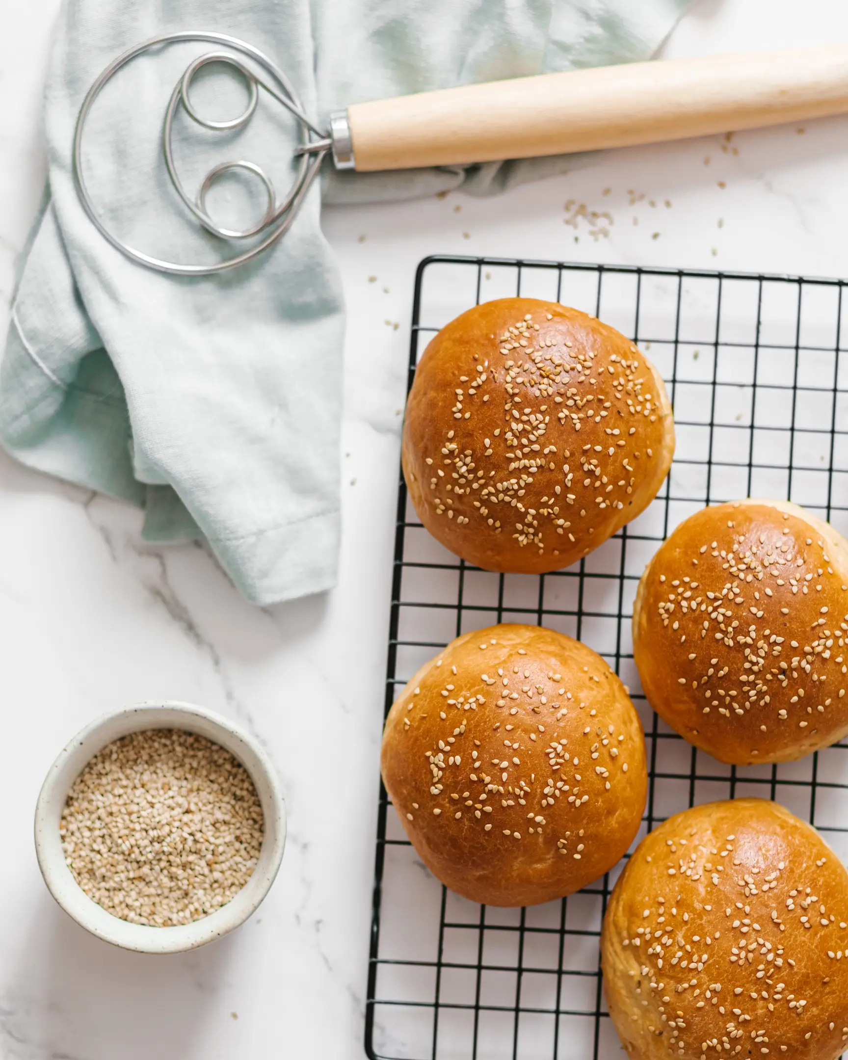There are burger buns on a pastry rack .  There are burger buns on a pastry rack to cool. On the side of them lies a tool for mixing dough and a towel. Below the frame are sesame seeds.