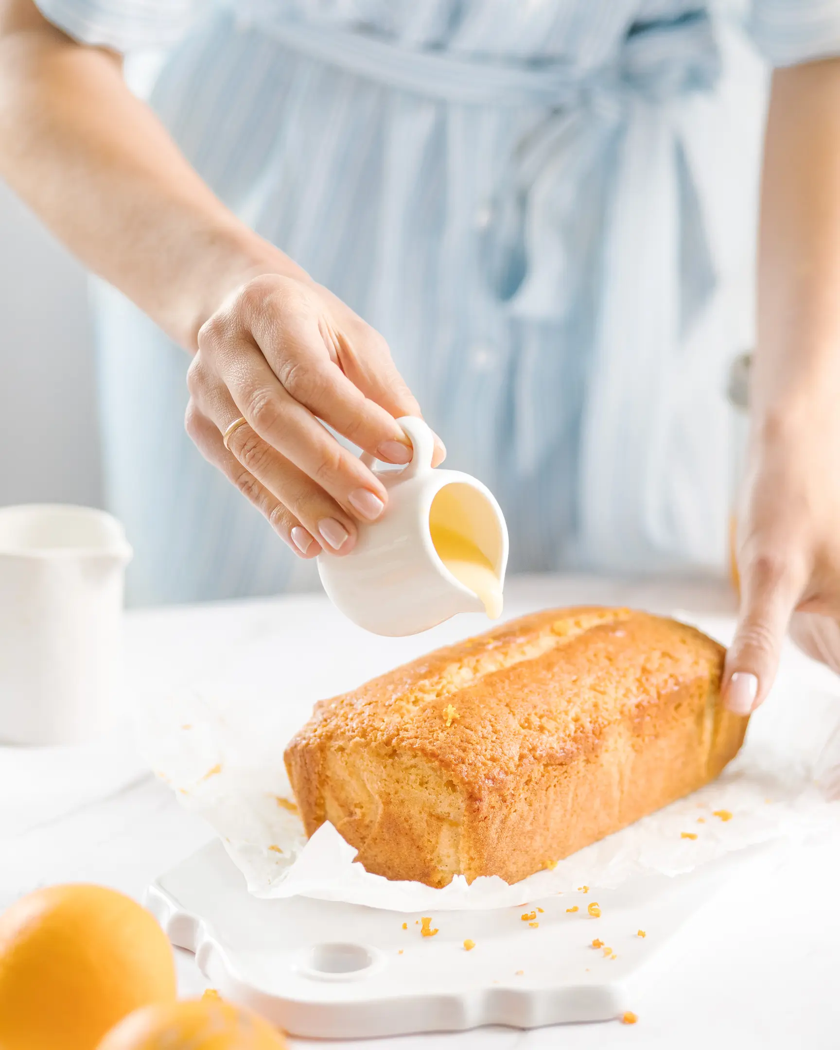 A woman pours orange icing on a cupcake.  A woman in a blue dress pours orange icing on a cupcake. The cupcake stands on a ceramic stand. Oranges are visible in the foreground, which emphasizes that the cupcake is made on the basis of oranges.