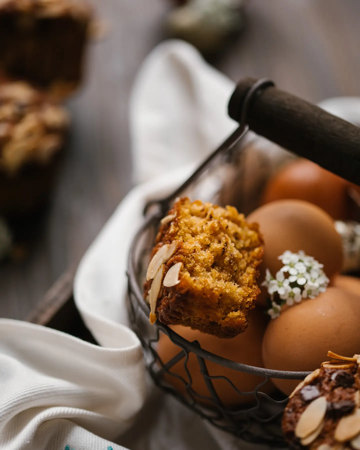 The cupcake is broken and its texture is visible. Chicken eggs are in a metal basket. Under the basket is an embroidered towel. There is a carrot cake in the basket. It is decorated with almond petals. The cupcake is broken and its texture is visible.