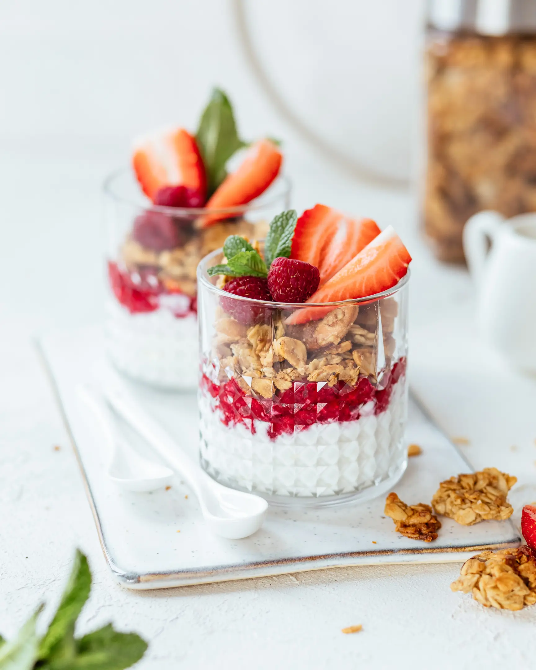 On a light background are 2 cups of yogurt and granola.  On a light background are 2 cups of yogurt and granola. They are filled with granola. The dessert is decorated with strawberries. In the background is a large can of granola.