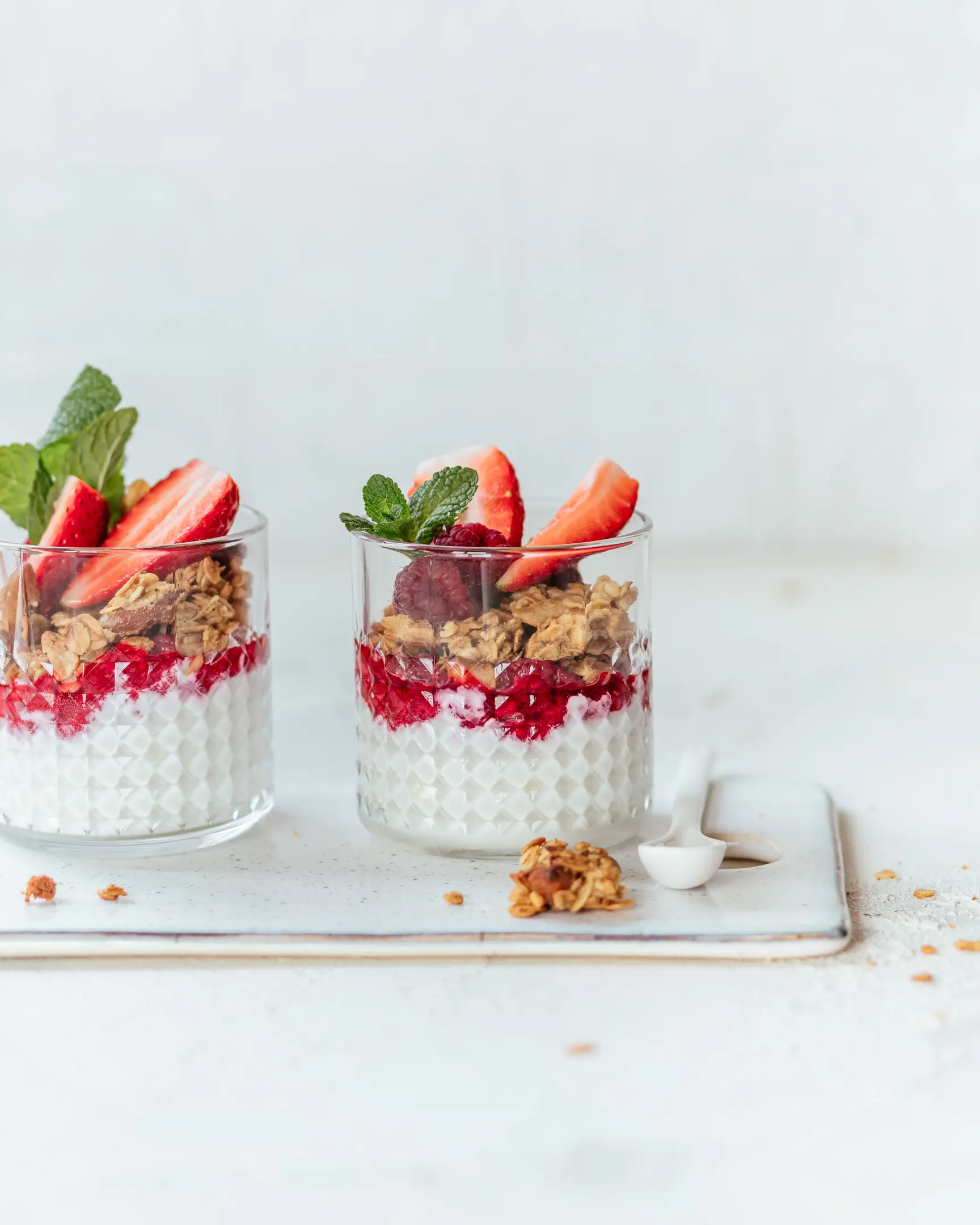 On a light background are 2 cups with granola and yogurt. On a light background are 2 cups of yogurt and granola. They are filled with granola. The dessert is decorated with strawberries. In the background is a large can of granola.