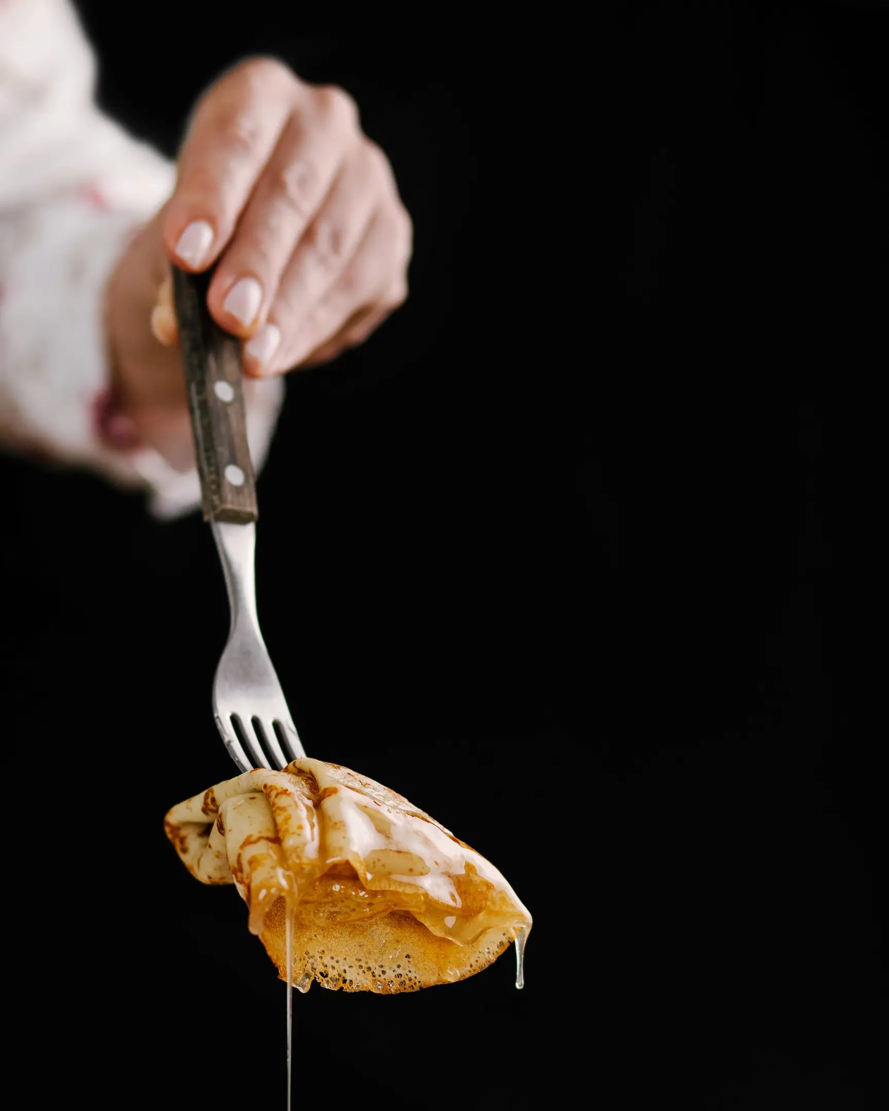A woman holds a fork in her hand, on which a pancake has been pricked. A woman holds a fork in her hand, on which a pancake has been pricked. The pancake is poured with honey. Honey drips from the pancake. Honey glitters in the rays of light.