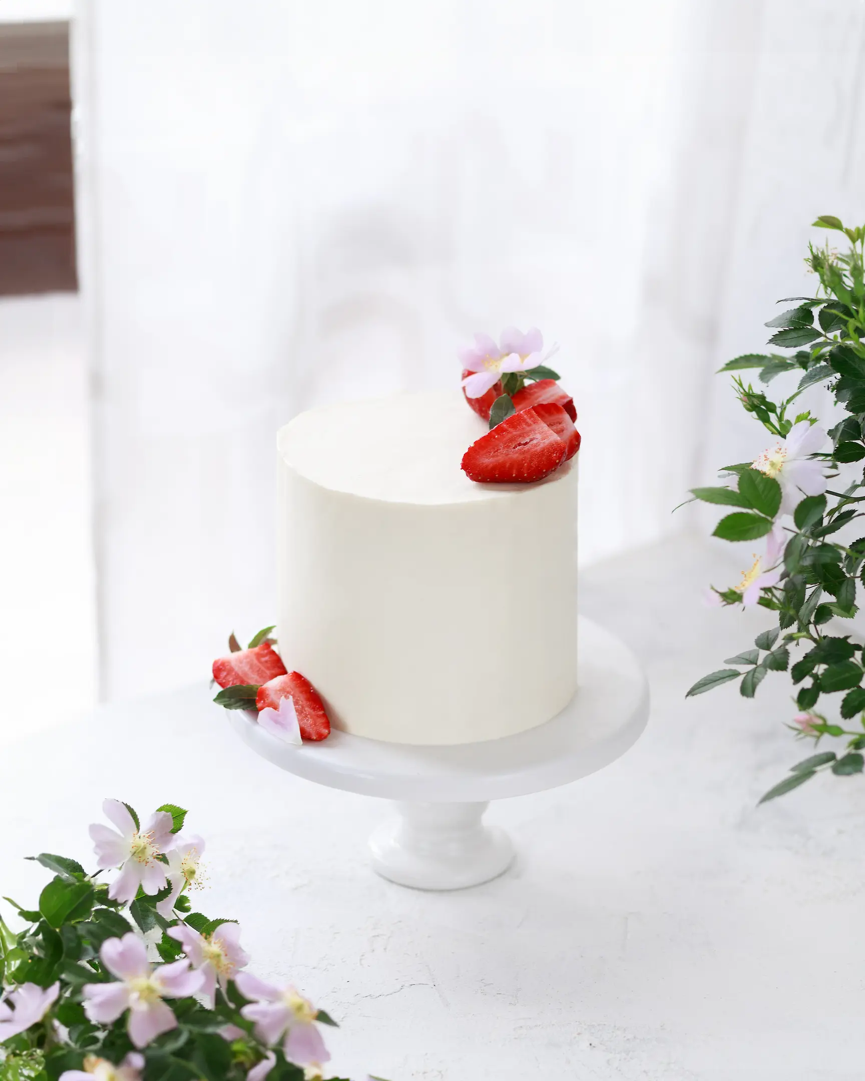 On a light background honey cake covered with white cream. On a light background opposite the window stands a cake decorated with berries. On the side of the frame, rosehip flowers are visible.