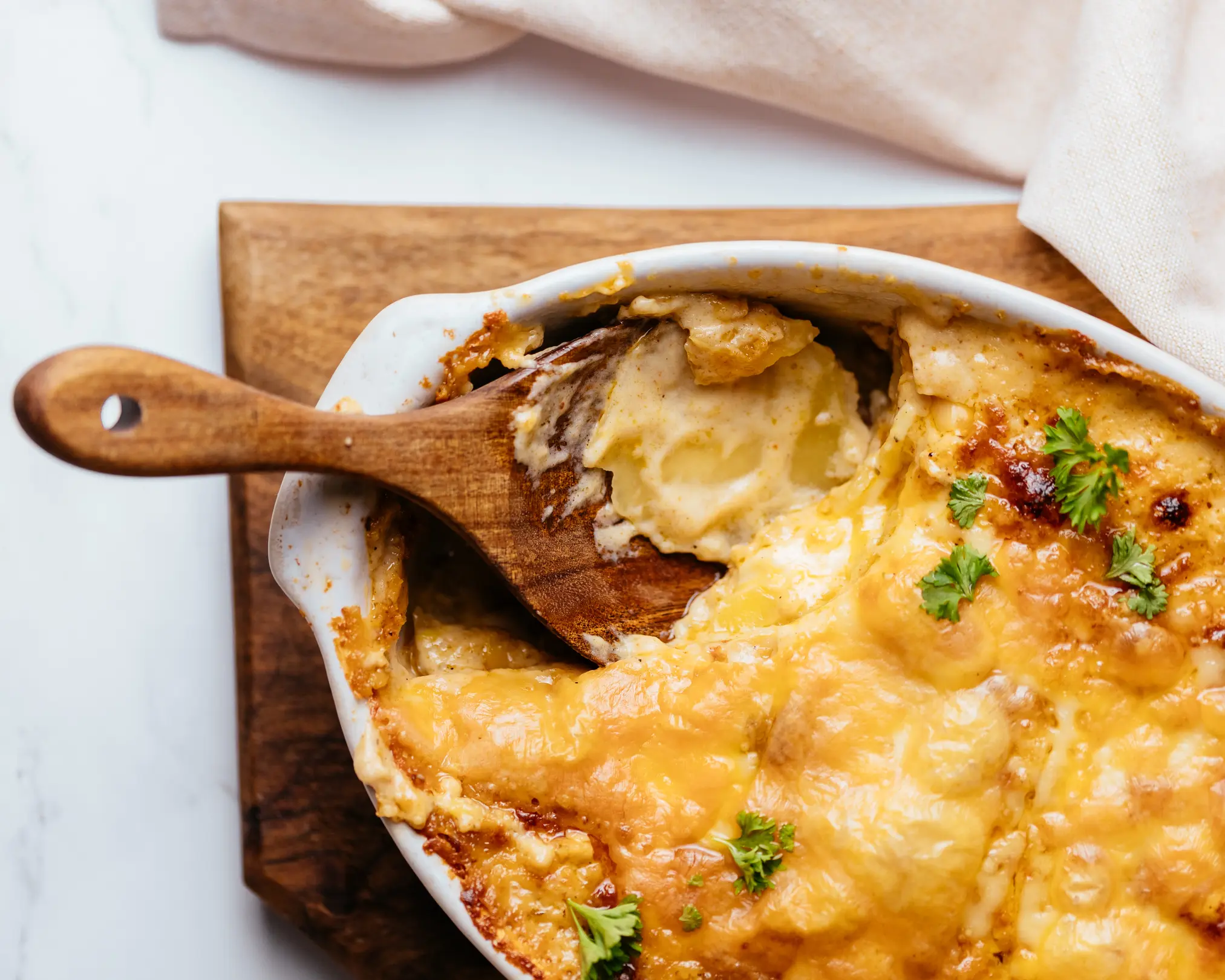 potato casserole with sauce. On a light background, there is a  wooden cutting board on it is an oval baking dish. It contains a potato casserole with sauce.