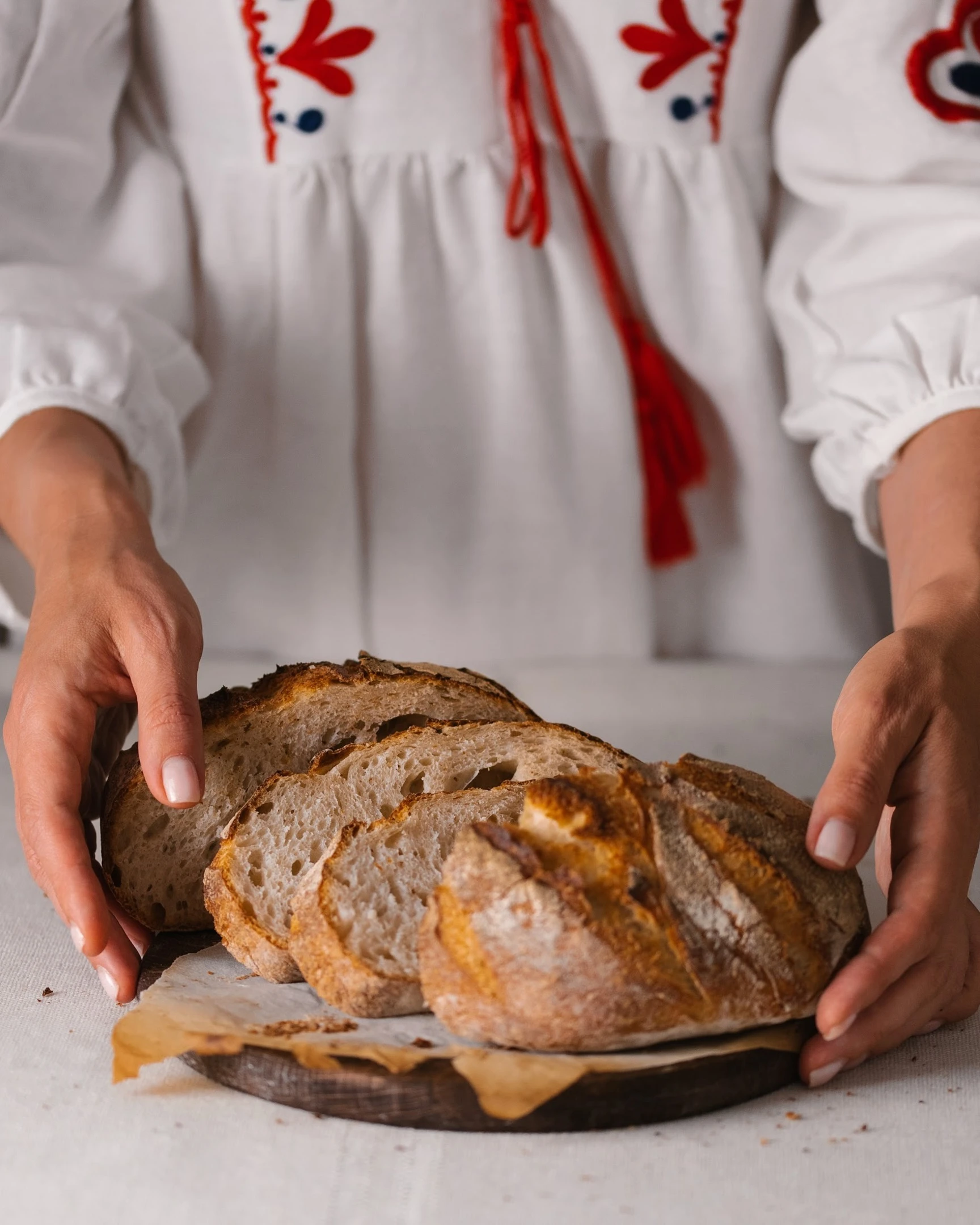 Sourdough bread   On the table lies a light linen tablecloth. On it is a wooden cutting board, and on the board is sliced bread. The woman is dressed in a dress with a red ornament.