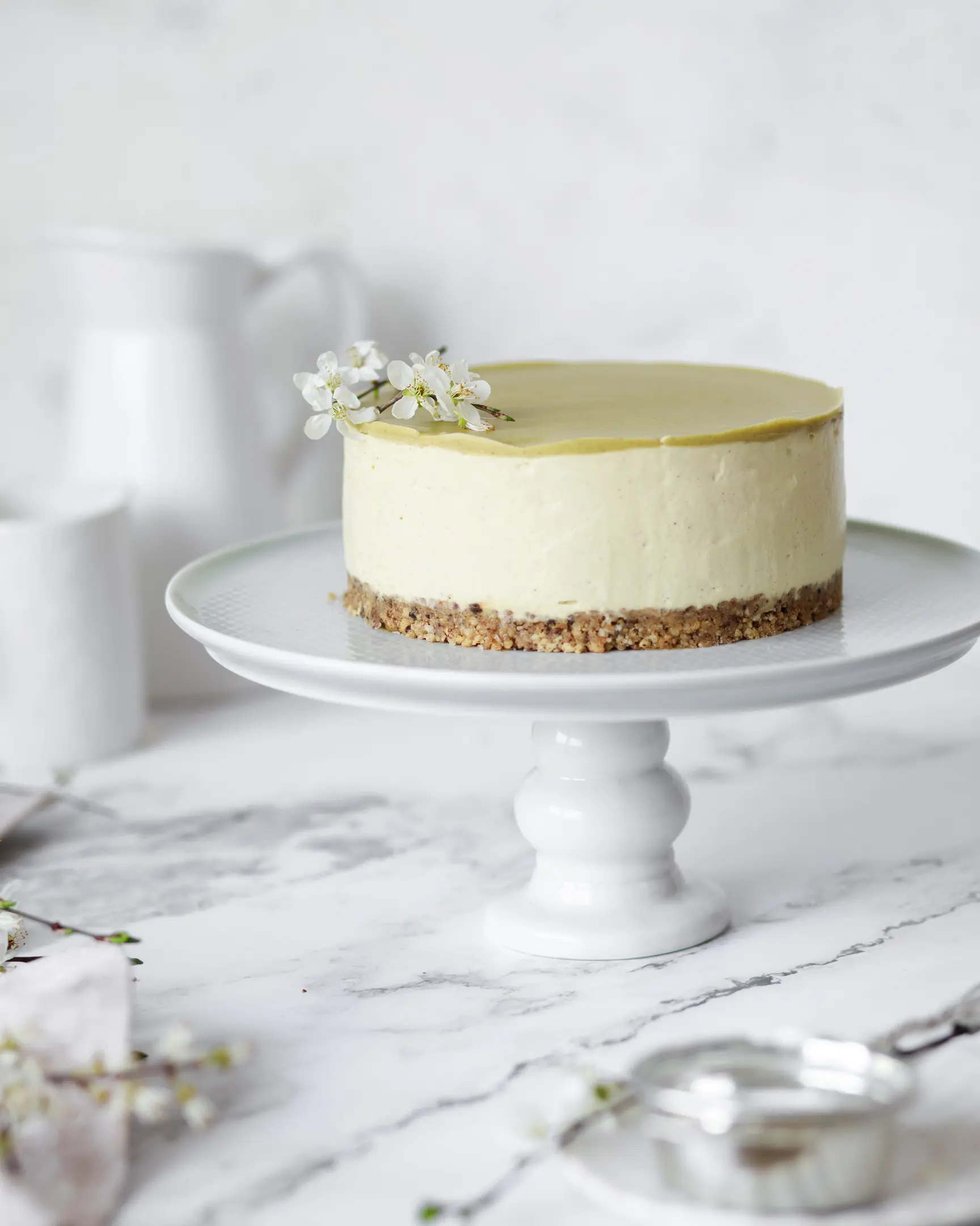 Сheesecake with pistachio paste  On a light background, on a high cake stand is a cheesecake with pistachio paste without baking. It is decorated with spring flowers.