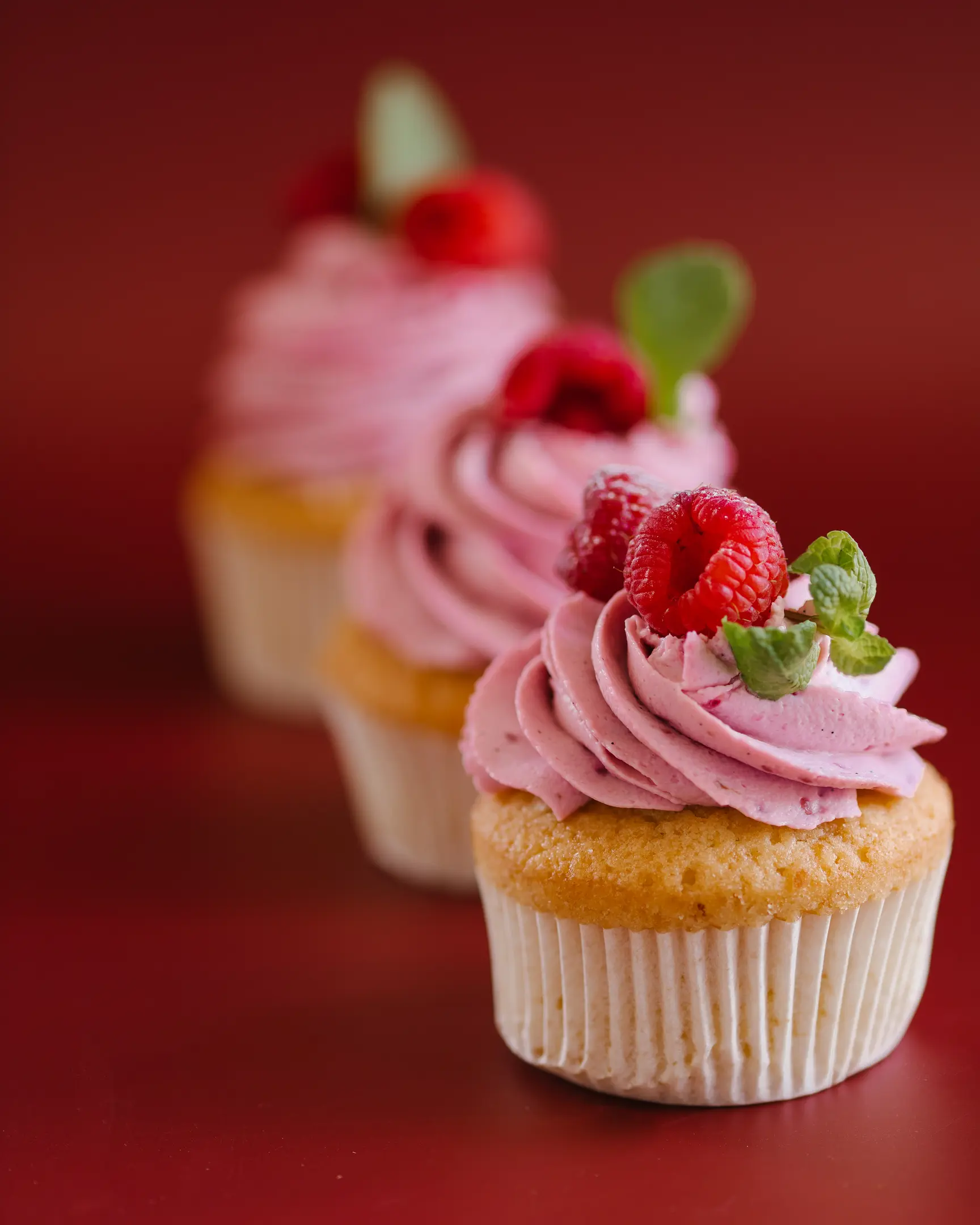 Сupcakes stand on a dark red background. 3 cupcakes stand on a dark red background. They are decorated with raspberry cream and raspberries.