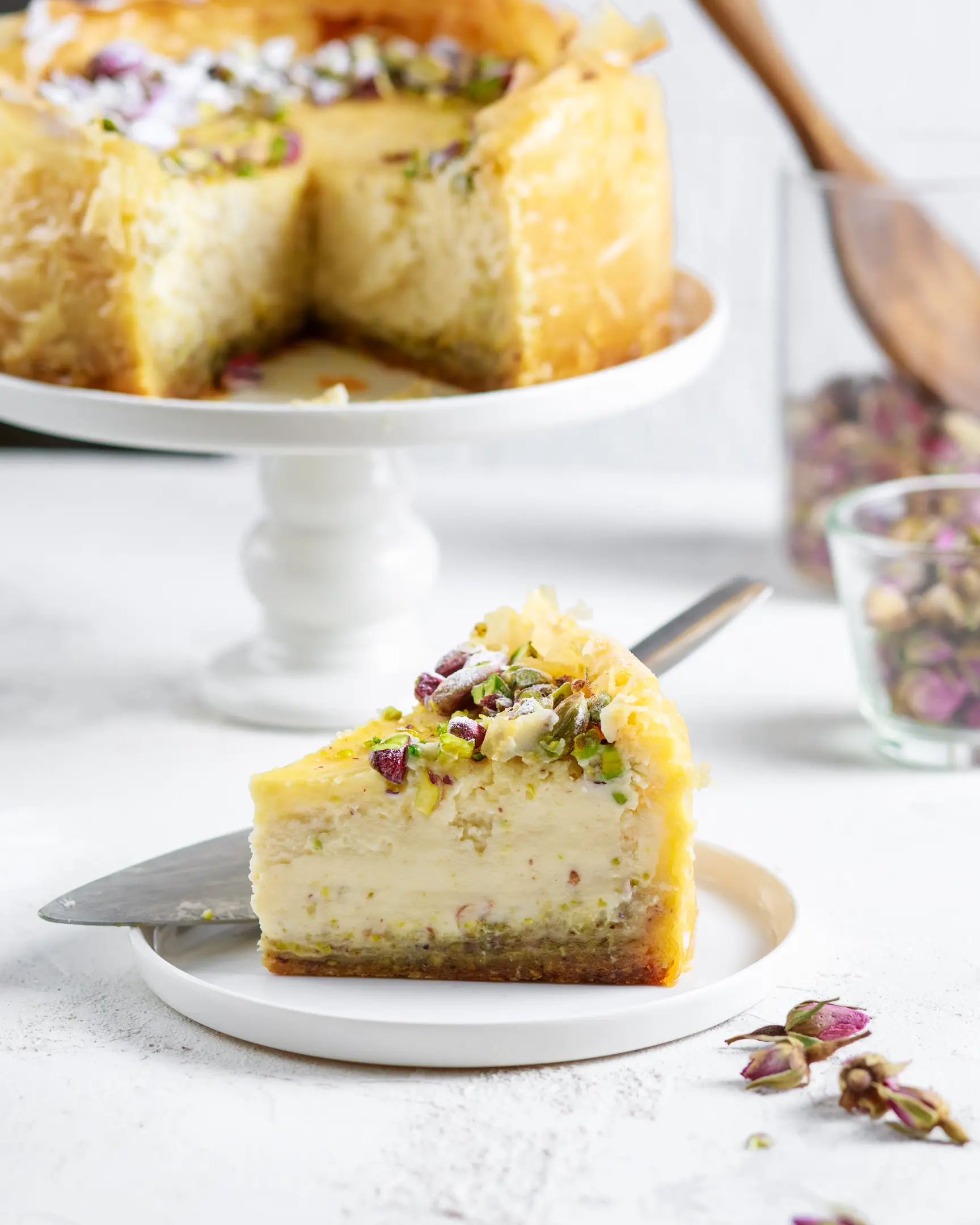 Baklava Cheesecake is standing on the cake stand.  There is a Baklava Cheesecake on the cake stand. A piece was cut off from it.   The cheesecake is decorated with pistachios and dried roses. In the foreground is a piece cut off from the general part. The cheesecake texture is visible.