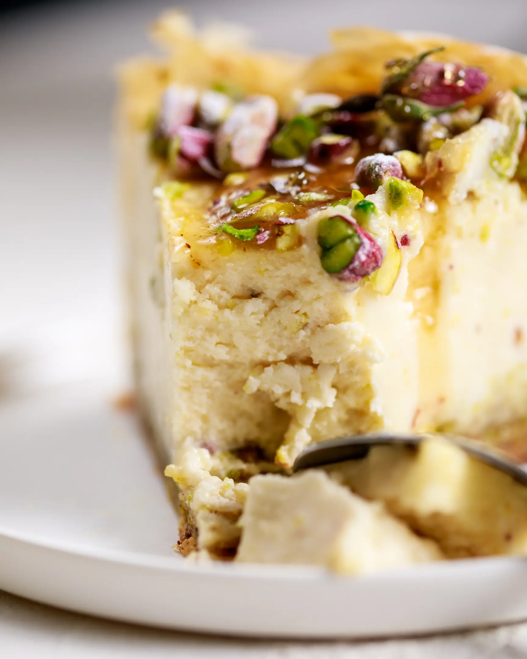 There is a piece of Baklava  cheesecake on the plate.  There is a piece of cheesecake on the plate. It is decorated with pistachio nuts. Cheesecake broken with a fork. The cheesecake texture is visible.