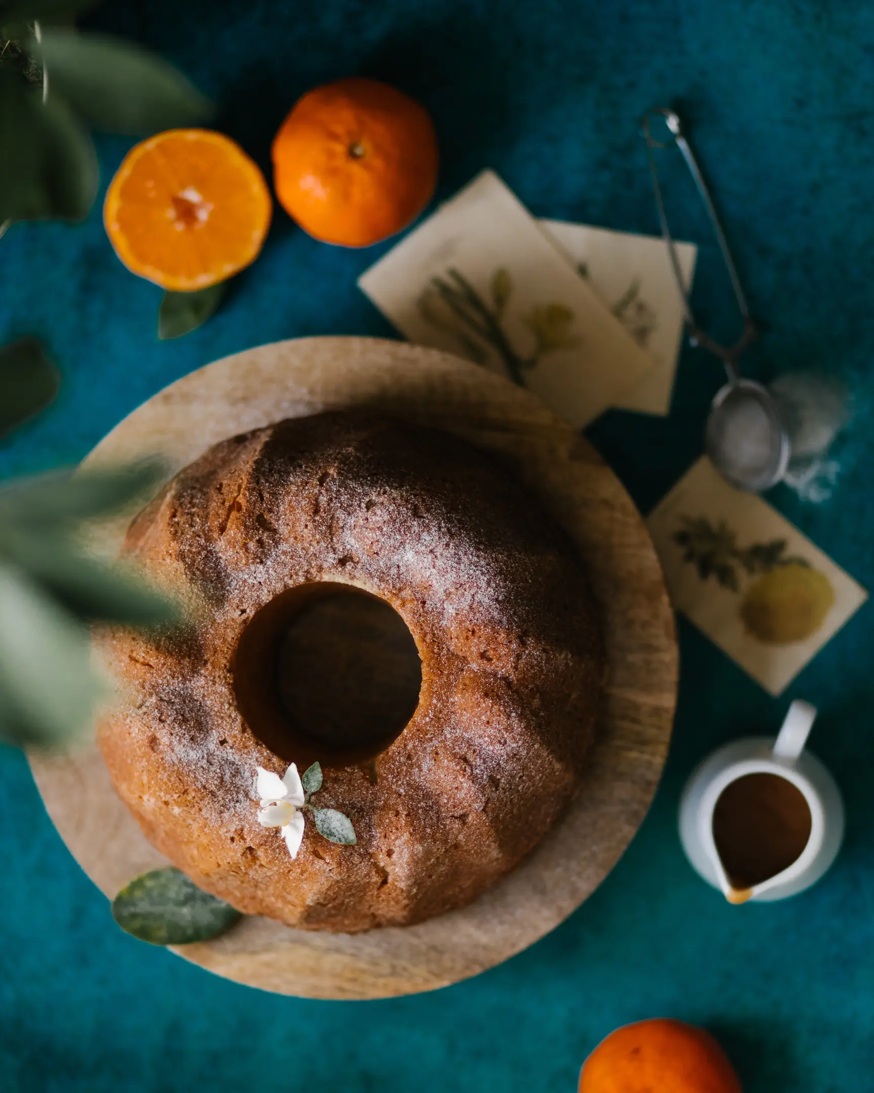 Кекс с мандарина. Поглед отгоре. Кекс с мандарина стои на ярък тюркоазен фон. Декорирана е с цитрусови цветя. Поглед отгоре. Кексът стои върху дървена тортена чиния. Около него има разпръснати листа с рисунки на цветя.