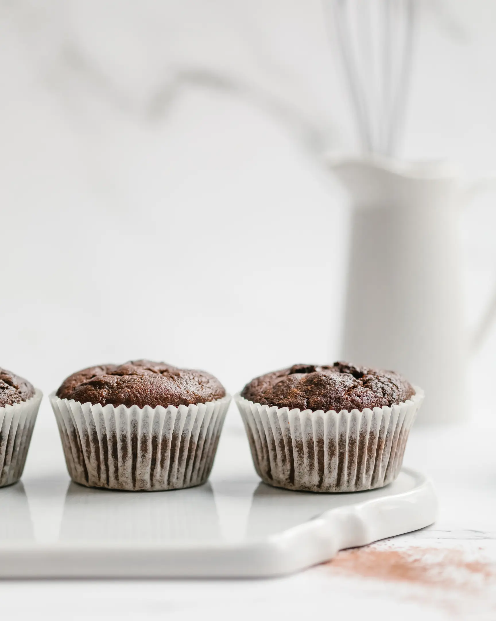 There are chocolate-colored cupcakes.  On a light table on a white ceramic cutting board there are chocolate-colored cupcakes. They are made without flour, eggs or refined sugar.