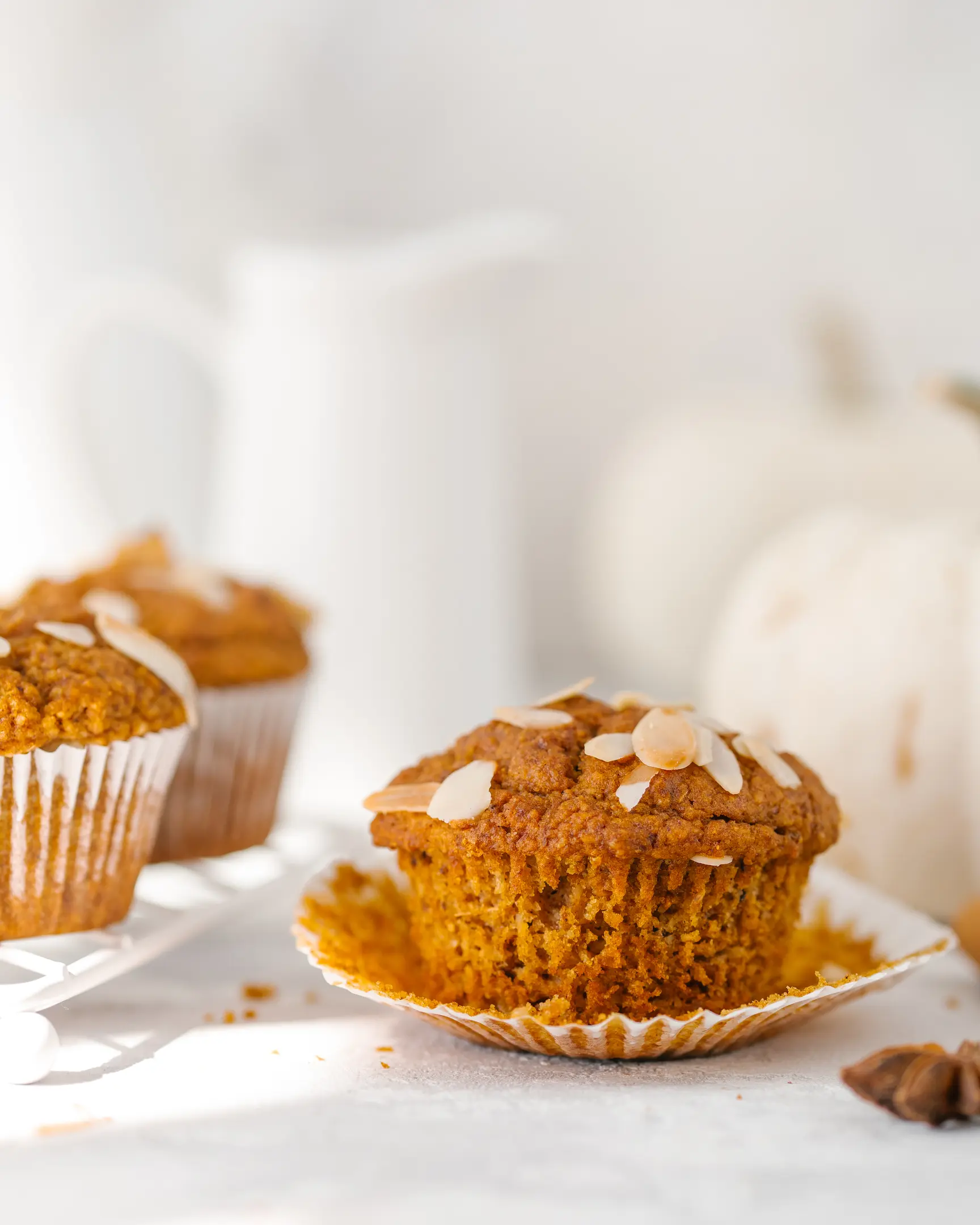 On a light table there is a sunny pumpkin muffin On a light table there is a sunny pumpkin muffin, it is decorated with slivered almonds. There are white pumpkins and a milk jug in the background.