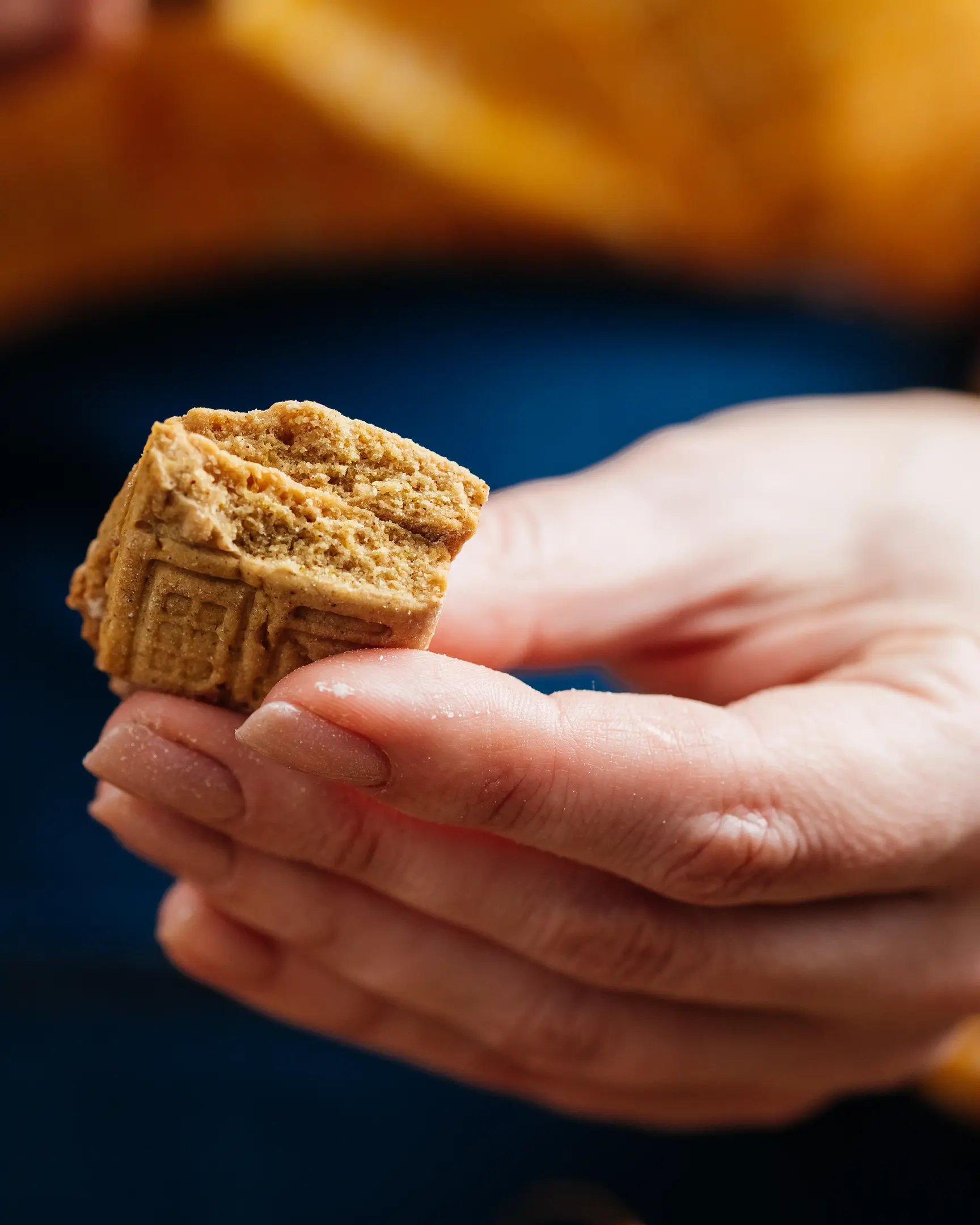 Cut of cookie The photo shows a close-up of a hand gently holding a beautifully detailed cookie in the shape of a house. The cookie features intricate imprints resembling windows, a door, and a roof, giving it the look of a charming, miniature home. The background is softly blurred, with warm yellow and blue tones, enhancing the cozy and inviting atmosphere. The cookie appears crisp and golden, suggesting it is freshly baked.