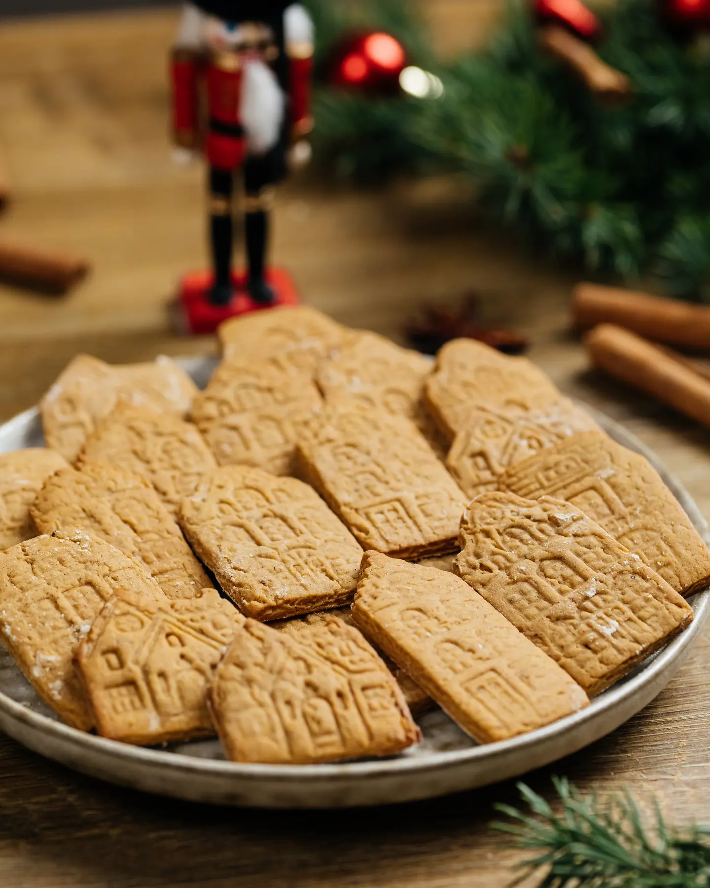 The photo shows a plate full of rectangular honey cookies, each featuring intricate designs that resemble old-fashioned European houses. The photo shows a plate full of rectangular honey cookies, each featuring intricate designs that resemble old-fashioned European houses. The cookies are golden-brown and neatly arranged on the plate, creating a cozy and festive atmosphere. In the slightly blurred background, there are cinnamon sticks, evergreen branches, red Christmas ornaments, and a wooden nutcracker figure, enhancing the holiday theme. The cookies look crispy, beautifully detailed, and perfect for a winter gathering or gift-giving.