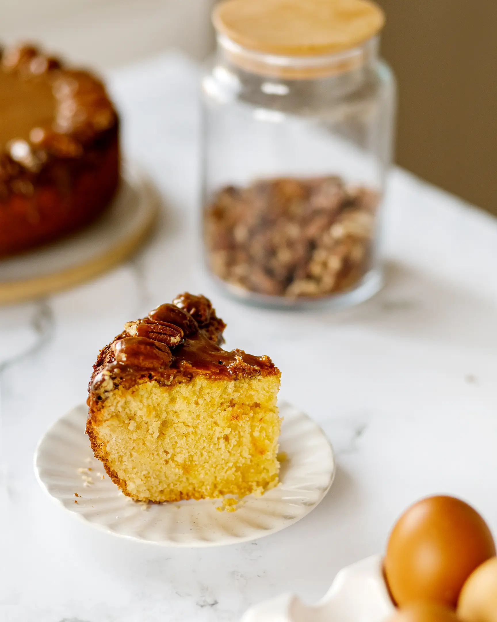 There is a piece of cut pecan cake on the table. There is a piece of cut pecan cake on the table. The cupcake is topped with caramel. In the background you can see a jar of nuts and the pie itself from which this piece was cut. In the foreground there are brown eggs in a light egg cup
