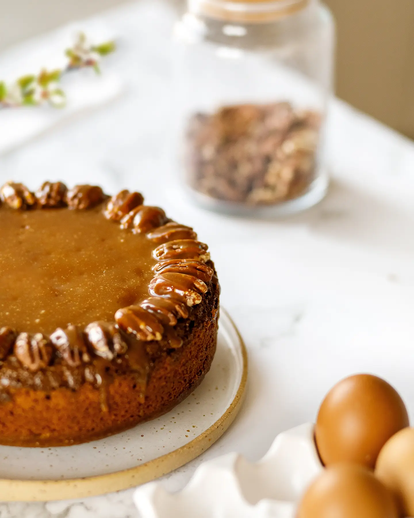 There is a   pecan cake on the table. There is a   pecan cake on the table. The cake is topped with caramel. In the background, you can see a jar of nuts. In the foreground there are brown eggs in a light egg cup