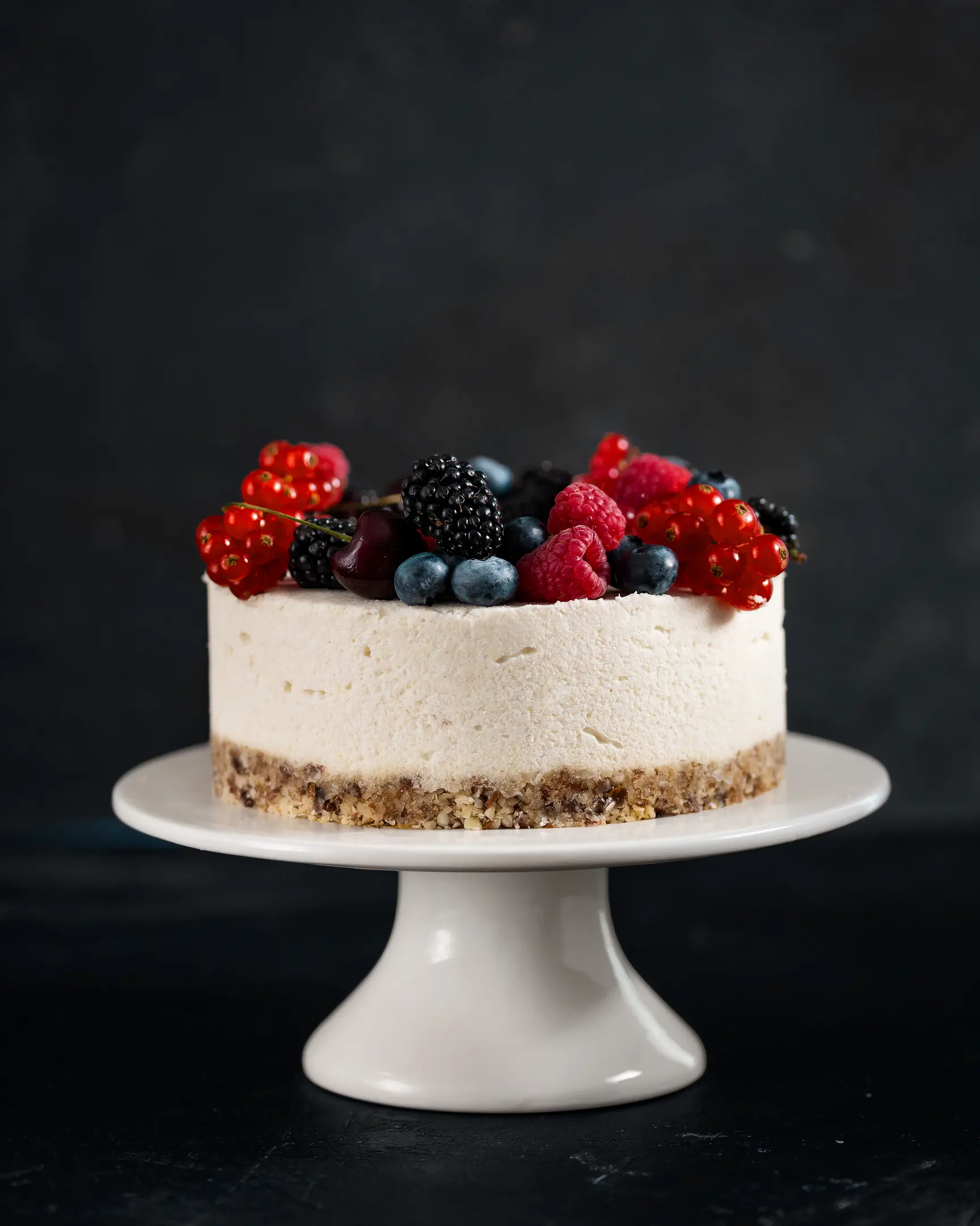 No Bake Coconut Cake on the dark Background The photo shows a beautifully presented cake on a white cake stand. The cake has a smooth, creamy white frosting coconut and is topped with an assortment of fresh berries. The berries include bright red currants, dark blackberries, blueberries, and raspberries, adding a burst of color and texture to the top of the cake. The cake's base appears to be a nutty or crumbly layer, contrasting with the creamy frosting above. The background is dark, which enhances the vibrant colors of the berries and the cake itself, making it the focal point of the image.