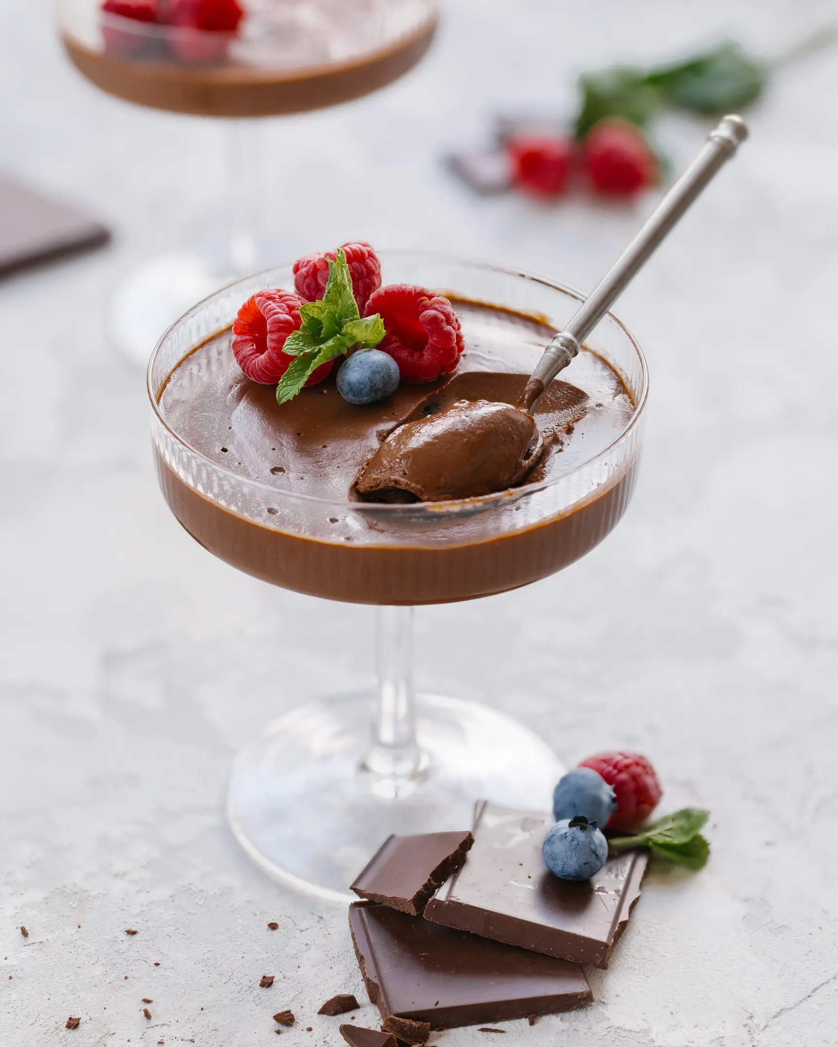 The photo shows a dessert in a tall-stemmed glass. It is a chocolate mousse. The photo shows a dessert in a tall-stemmed glass. It is a chocolate mousse. The mousse has a smooth and creamy texture, with an imprint from a spoon that is dipped into the dessert and holding a small portion. On the surface of the mousse, there are several fresh raspberries and blueberries, as well as a sprig of fresh mint. Next to the glass, on the table, there are pieces of dark chocolate and a few berries, complementing the overall composition. The background of the photo is light and blurred, emphasizing the focus on the dessert.