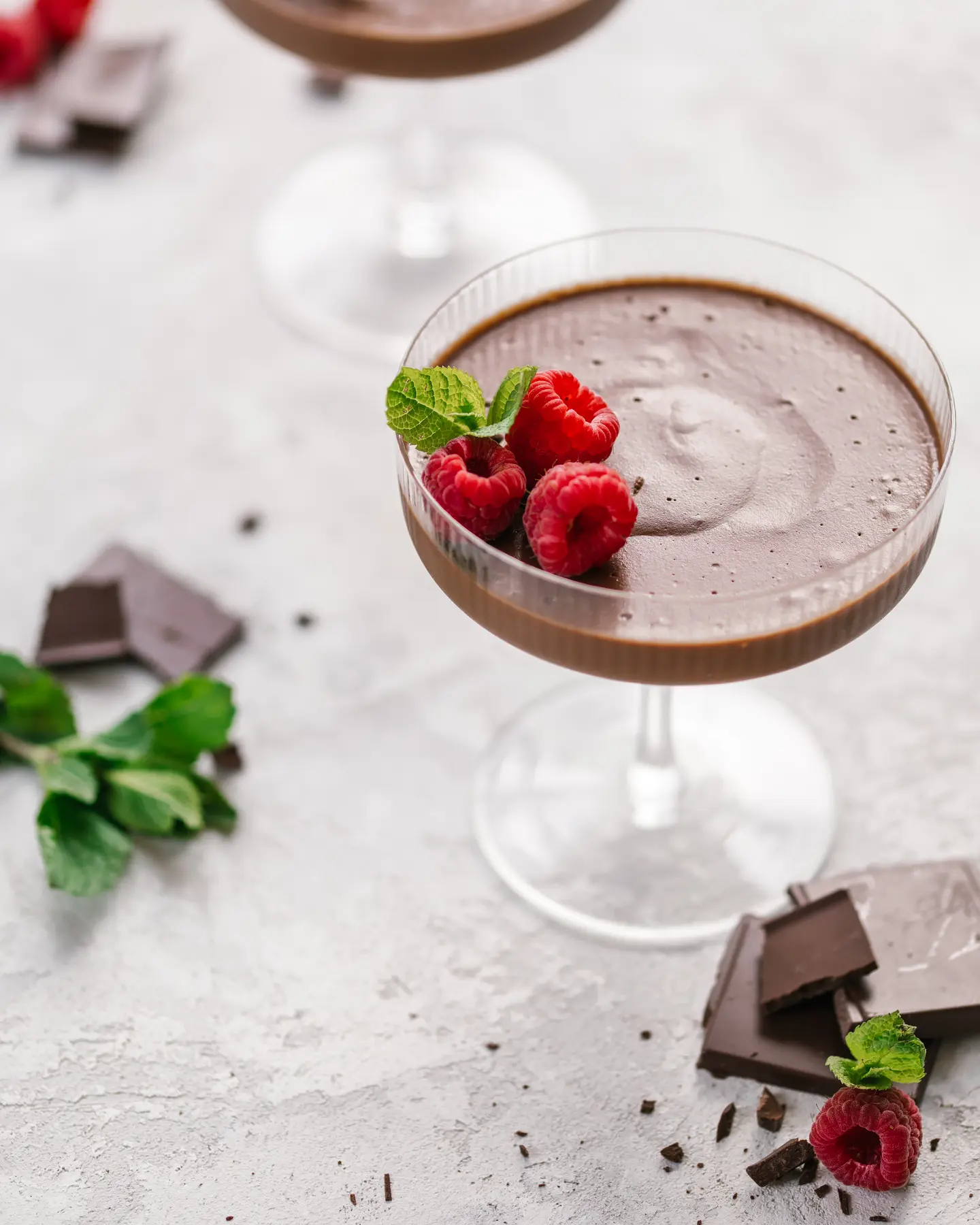 It is a chocolate mousse  in a tall-stemmed glass. The photo shows a dessert in a tall-stemmed glass. It is a chocolate mousse.  On the surface of the mousse, there are several fresh raspberries. Next to the glass, on the table, there are pieces of dark chocolate and a few berries, complementing the overall composition. The background of the photo is light and blurred, emphasizing the focus on the dessert.