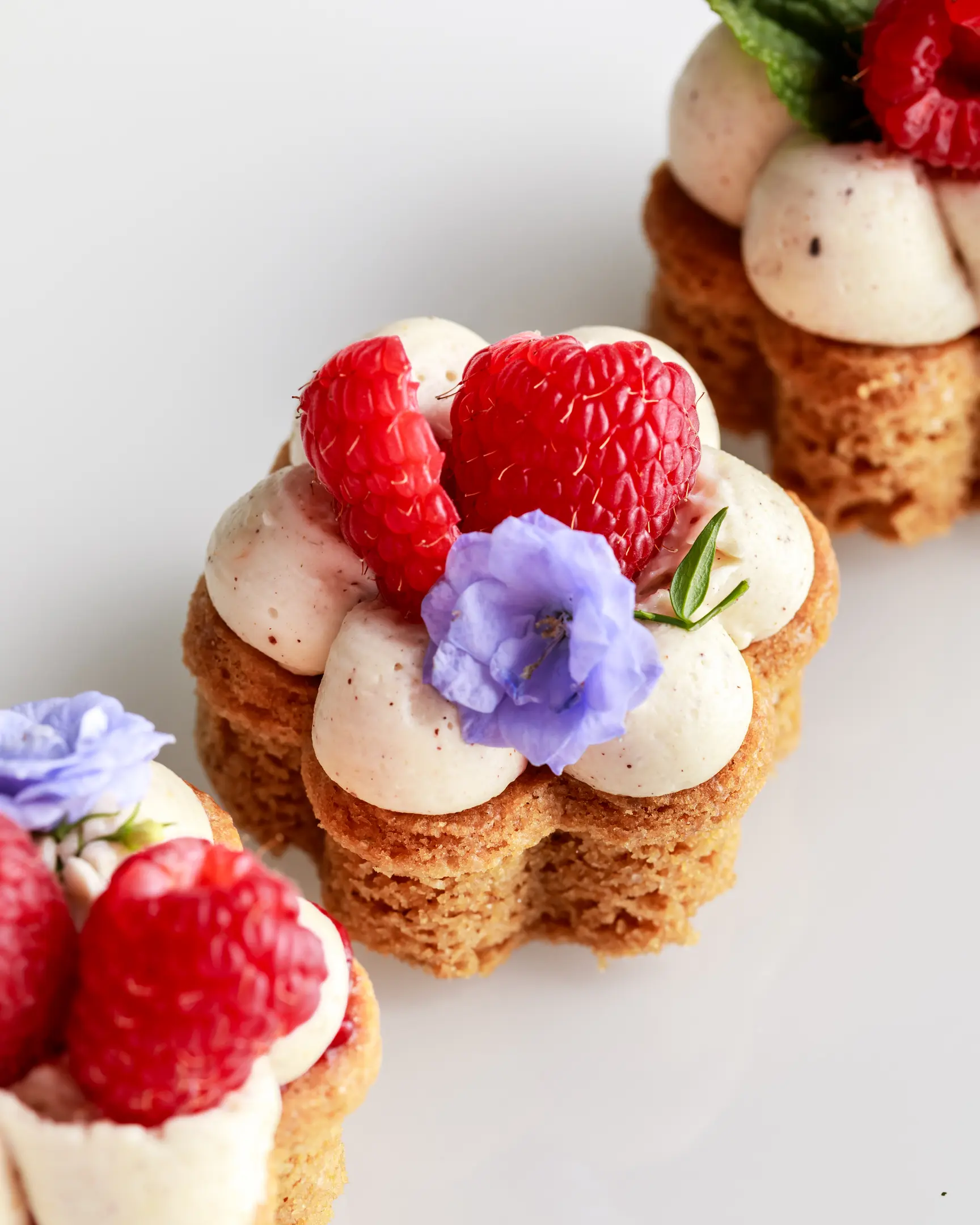 Close-up photo of three beautifully decorated, flower-shaped cookies. This photo showcases a close-up of three beautifully decorated, flower-shaped cookies. Each cookie has a golden-brown, textured base, resembling a delicate flower with six rounded petals. Atop each petal sits a generous dollop of smooth, creamy nut-flavored ganache, creating a visually pleasing contrast with the darker cookie base. In the center of each flower, there are fresh, plump raspberries, adding a vibrant red pop of color. The cookies are further adorned with small, delicate, purple edible flowers, and a few tiny green leaves, enhancing their elegant and refined appearance.