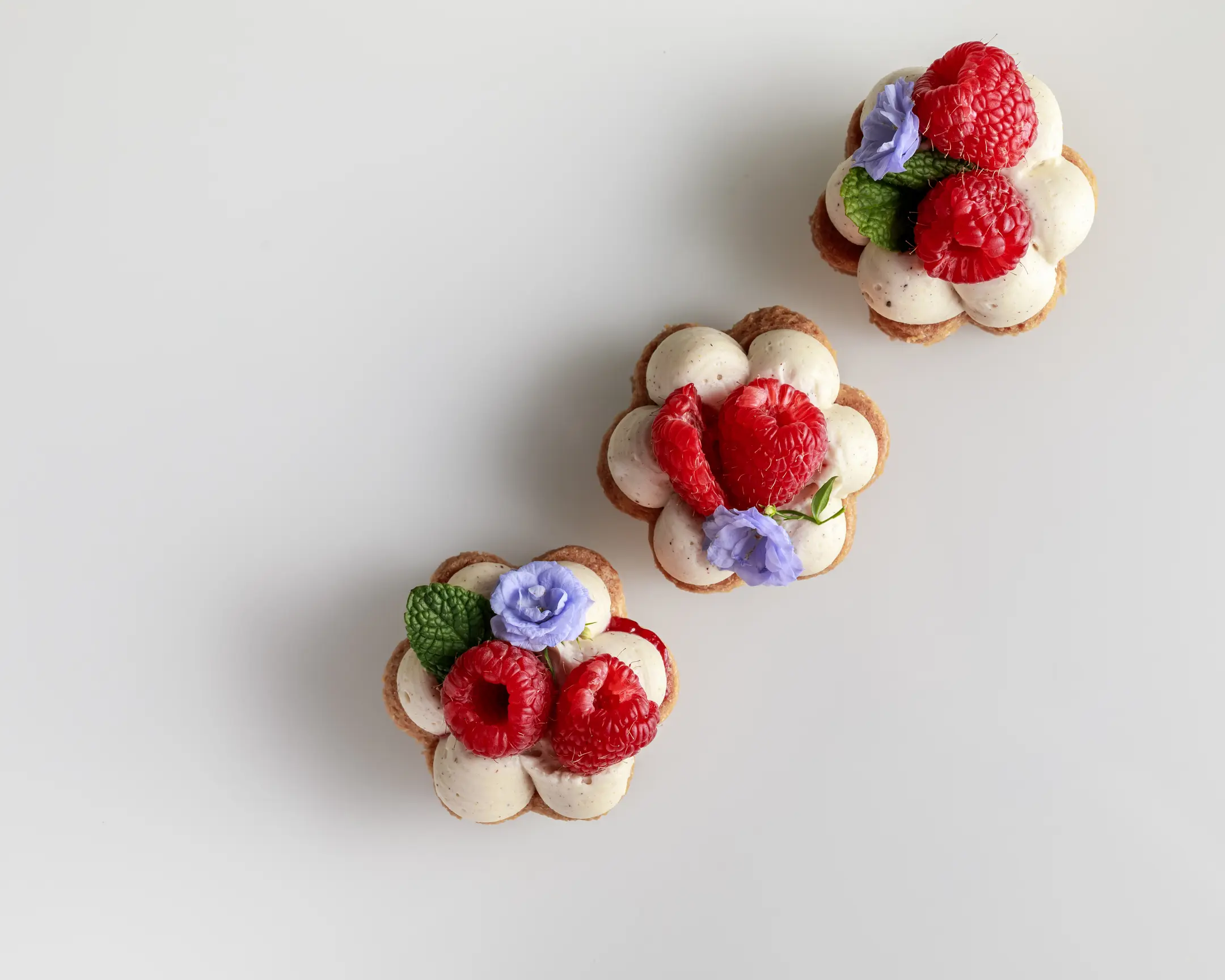 This photo showcases a view from above of three beautifully decorated, flower-shaped cookies.  This photo showcases a view from above of three beautifully decorated, flower-shaped cookies. Each cookie has a golden-brown, textured base, resembling a delicate flower with six rounded petals. Atop each petal sits a generous dollop of smooth, creamy nut-flavored ganache, creating a visually pleasing contrast with the darker cookie base. In the center of each flower, there are fresh, plump raspberries, adding a vibrant red pop of color. The cookies are further adorned with small, delicate, purple edible flowers, and a few tiny green leaves, enhancing their elegant and refined appearance.