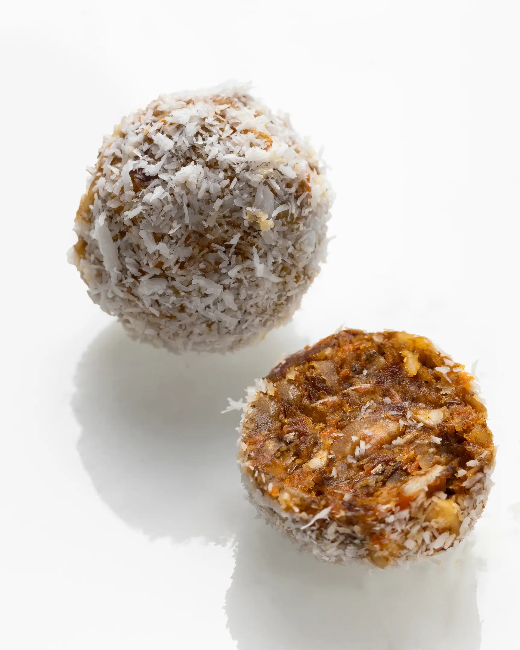 Two round candies covered with coconut flakes In the photo, there are two round candies covered with coconut flakes. One is whole, and the other is cut in half to reveal its internal texture. Inside, you can see a dense orange-colored mixture with bits of nuts and other natural ingredients. The candies look like a homemade or natural dessert made from carrots, nuts, and dried fruits, without added sugar.