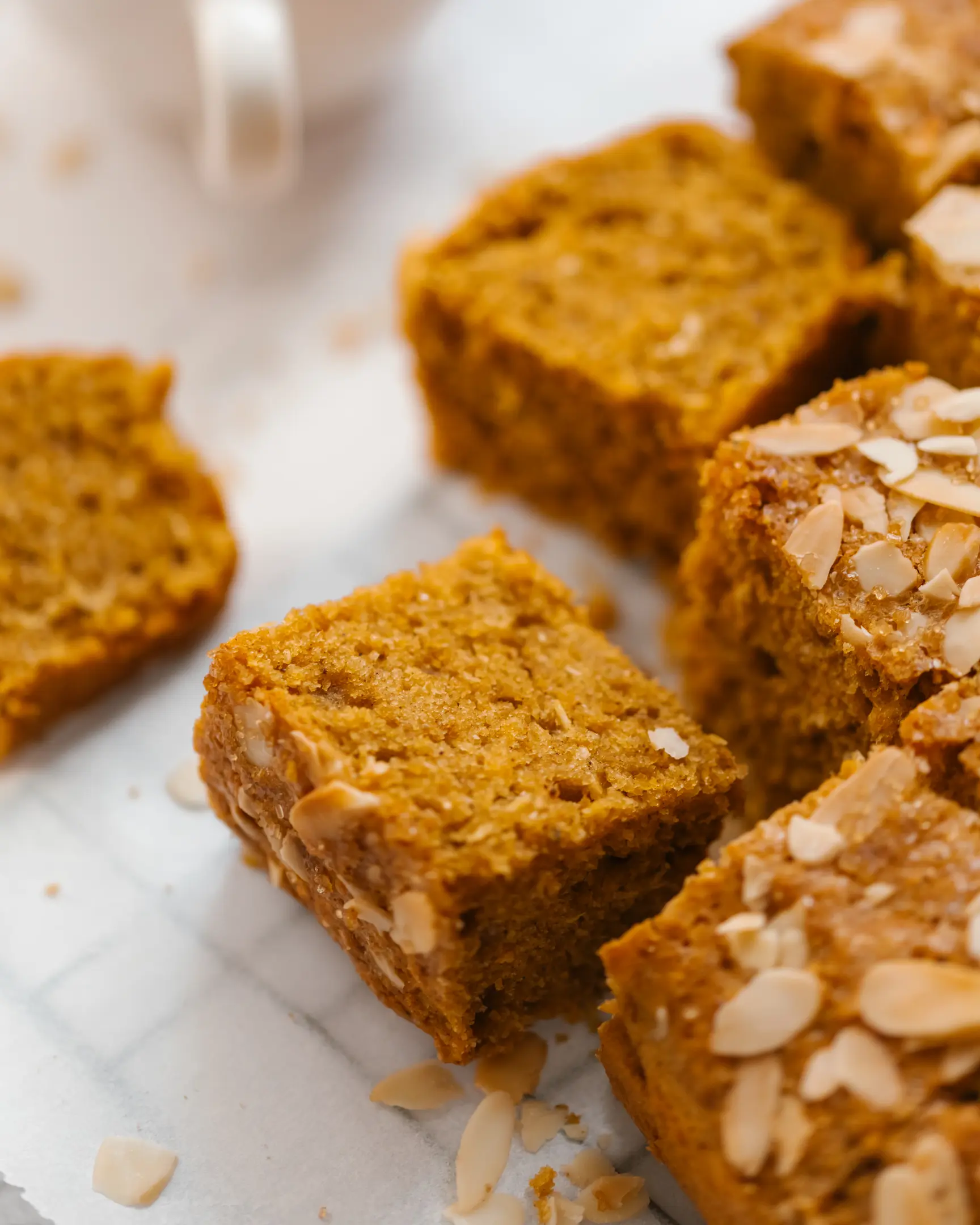 Golden-brown cake squares, arranged closely together on a white surface. The photo shows several pieces of golden-brown cake squares, arranged closely together on a white surface. The texture of the cake looks soft and moist, with visible almond flakes scattered on top of some pieces for decoration. The cakes have a warm, rich color, likely from the inclusion of corn and spices, giving them an inviting and homemade appearance. The focus is on the cakes, highlighting their fluffy interior and slightly crumbly edges, making them look delicious and perfect for autumn.