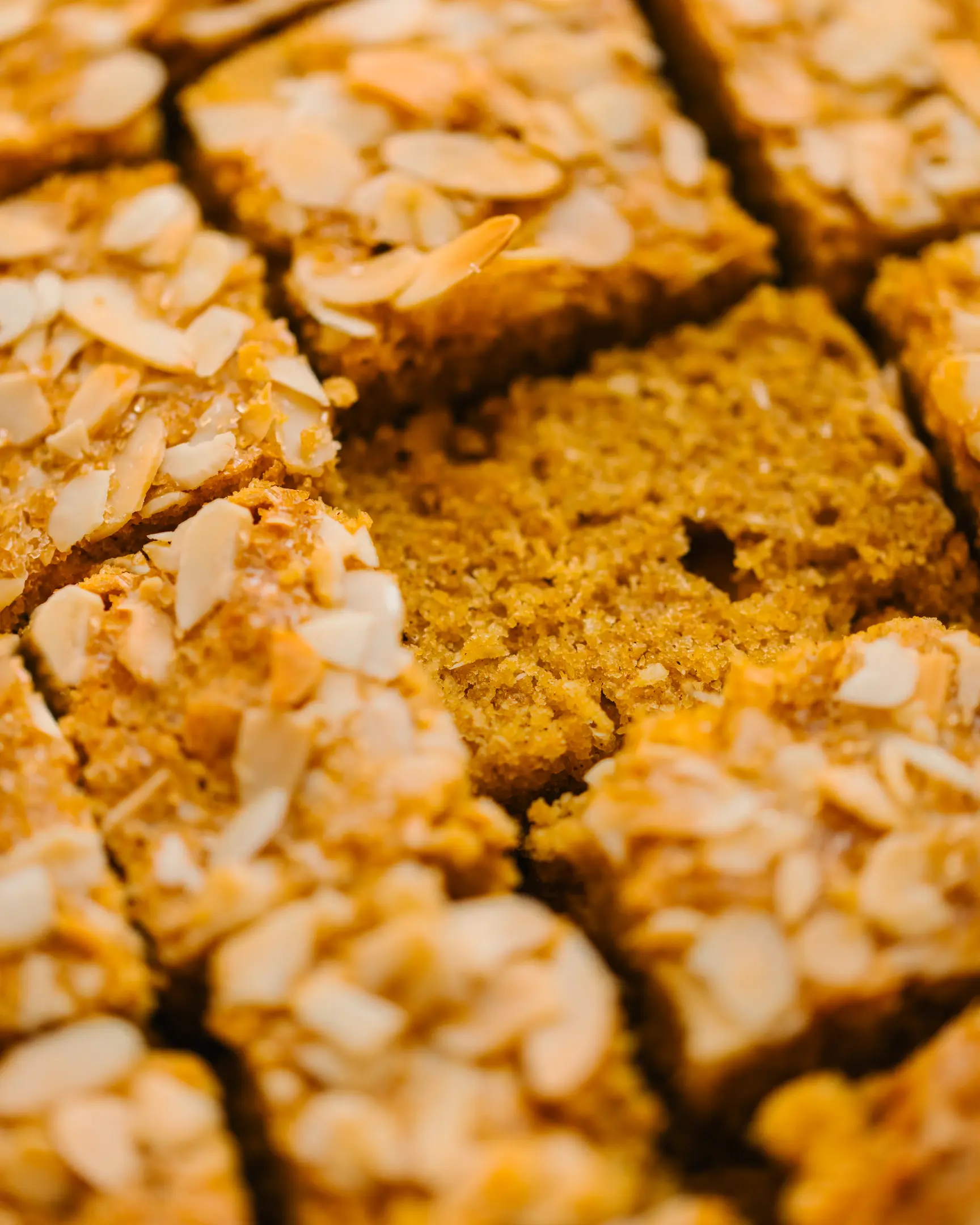  close-up pieces of a golden-brown cake cut into squares The photo shows close-up pieces of a golden-brown cake cut into squares. The cake has a moist and fluffy texture, with a warm orange hue, likely due to corn and spices used in the recipe. The surface is decorated with thin almond flakes that add a crunchy topping. One square in the center is slightly removed, revealing the soft interior of the cake. The overall presentation highlights the homemade and inviting nature of the dessert, making it look perfect for sharing during autumn or as a comforting treat.