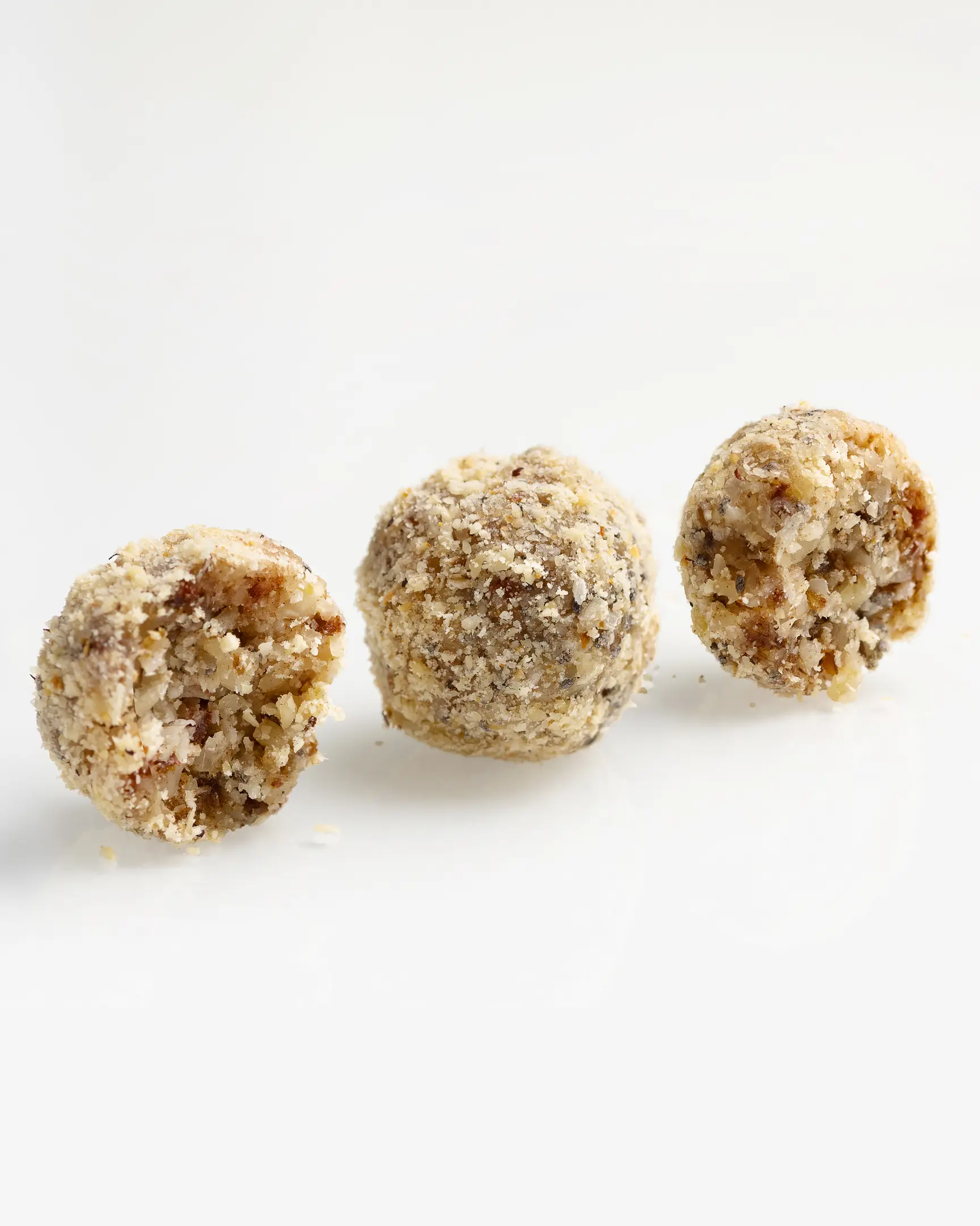 Three small round candies arranged in a row on a light background. The photo shows three small round candies arranged in a row on a light background. They are coated with a crumbly layer of finely chopped nuts or breadcrumbs, giving them a textured and slightly rough appearance. The candies look homemade, with an uneven, natural surface. Inside, you can see bits of ingredients that resemble a mix of nuts and dried fruits, creating a dense, slightly grainy texture. The colors of the candies are warm, with shades of golden, brown, and creamy tones, suggesting the use of natural ingredients like almonds and dried apricots.