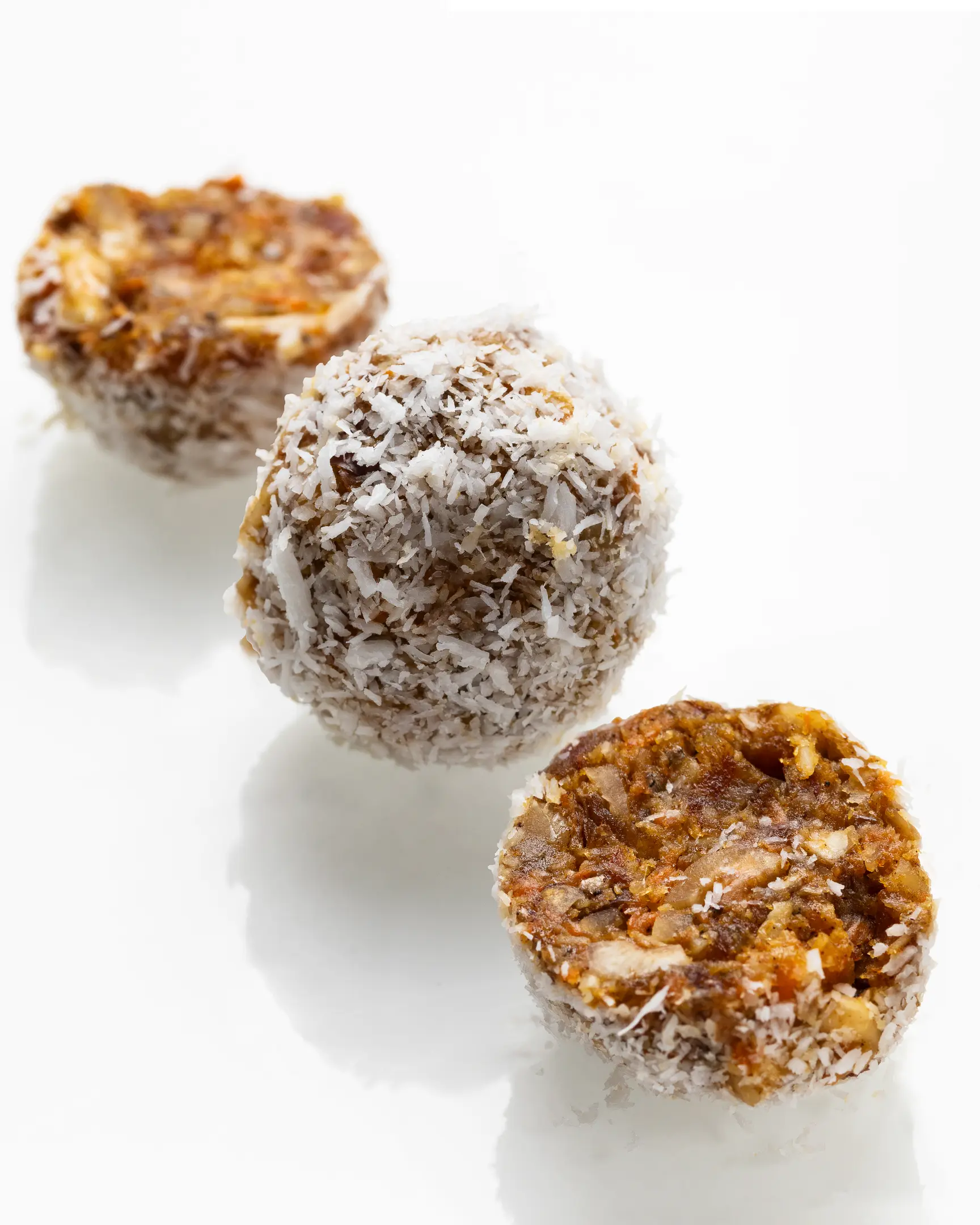 small round carrot  candies arranged in a diagonal The photo shows three small round carrot candies arranged in a diagonal on a light background. They are coated with a crumbly layer of finely chopped nuts or breadcrumbs, giving them a textured and slightly rough appearance. The candies look homemade, with an uneven, natural surface. Inside, you can see bits of ingredients that resemble a mix of nuts and dried fruits, creating a dense, slightly grainy texture. The colors of the candies are warm, with shades of golden, brown, and creamy tones, suggesting the use of natural ingredients like almonds and dried apricots.