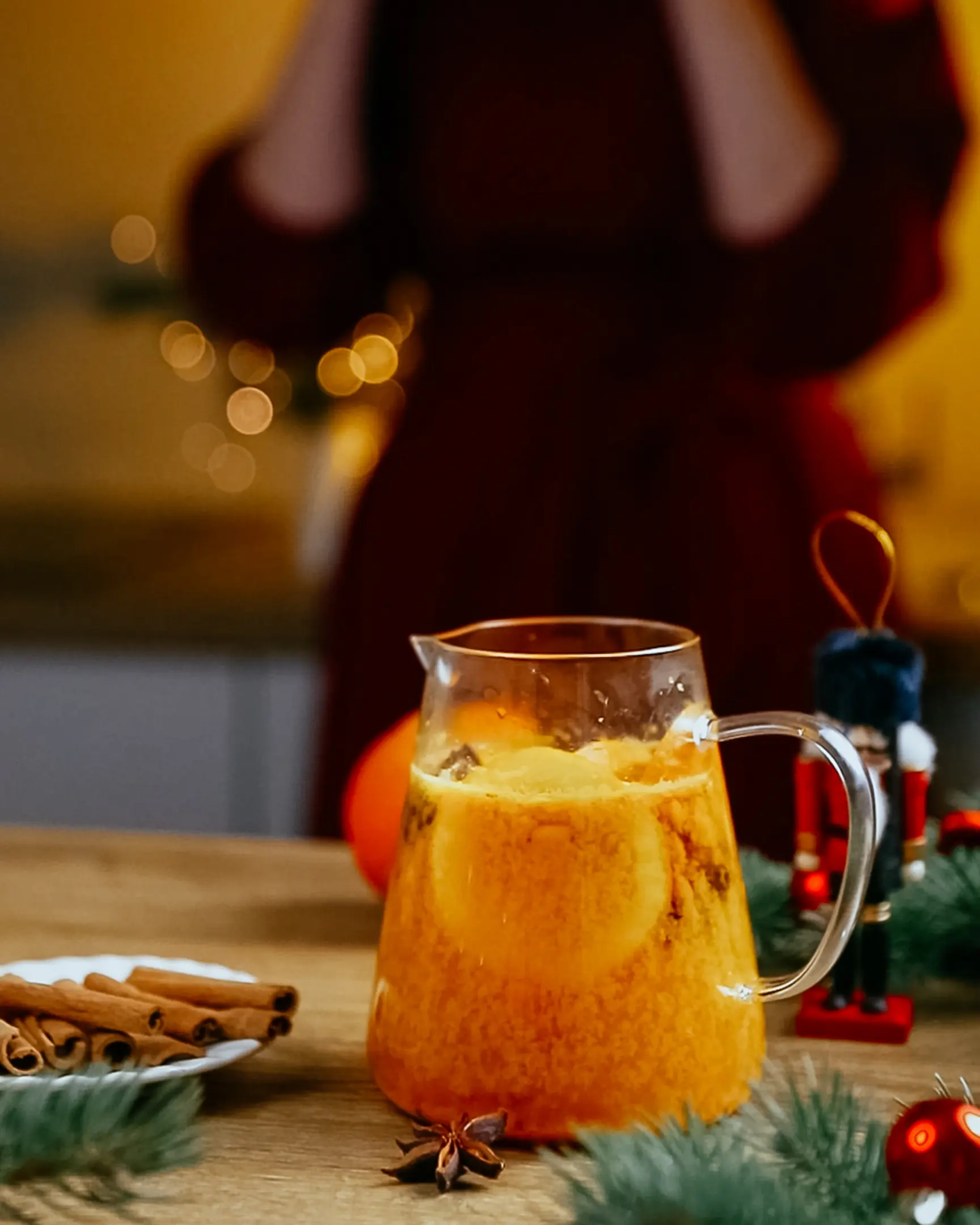 The orange tea made from sea buckthorn. The image shows a cozy, warm scene in a kitchen or dining area. In the foreground, there is a transparent glass jug filled with a bright orange tea made from sea buckthorn, slices of orange, and visible spices such as cinnamon sticks and star anise. The tea looks rich and inviting, with floating ingredients that enhance its texture and color. To the left, there is a small plate with cinnamon sticks, and evergreen branches frame the scene, adding a festive touch. In the background, out of focus, a person in a red dress is holding a cup near their face, as though sipping the tea, with warm, soft lighting creating a comforting and homely atmosphere.