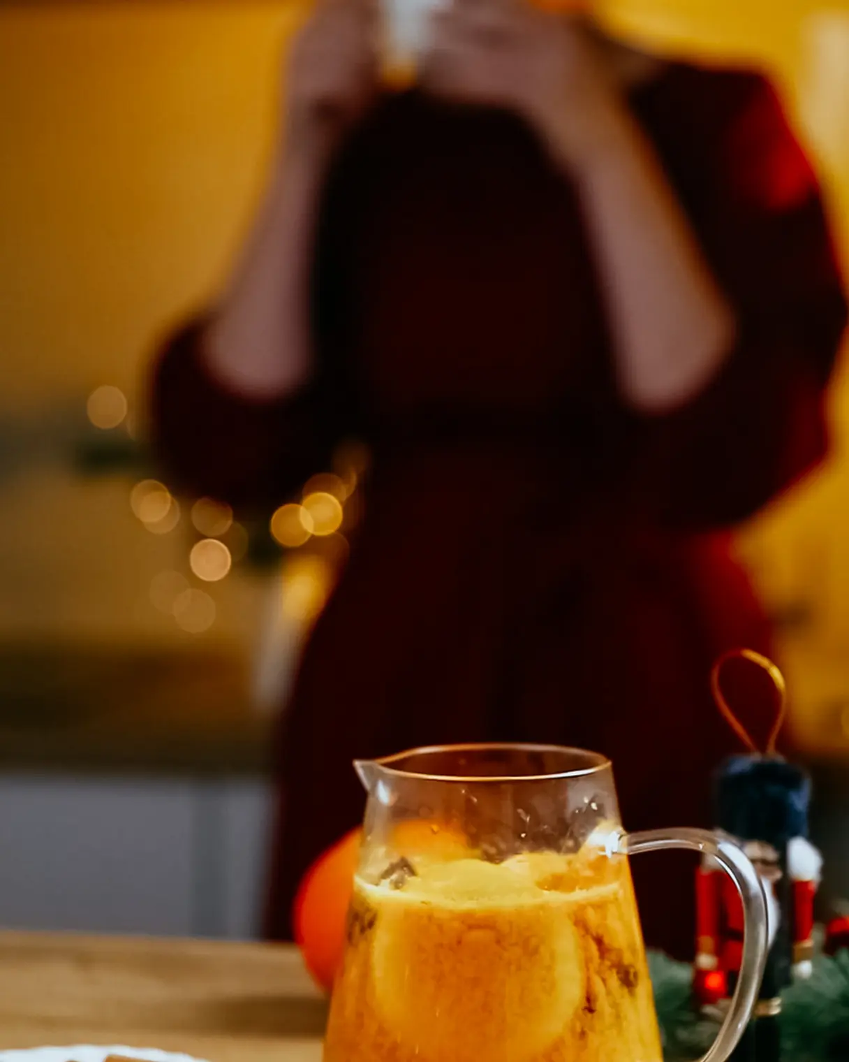 A woman in a burgundy dress against an orange-brown background drinks sea buckthorn tea from a white cup.  A woman in a burgundy dress against an orange-brown background drinks sea buckthorn tea from a white cup. In the foreground there is a teapot and Christmas decorations.