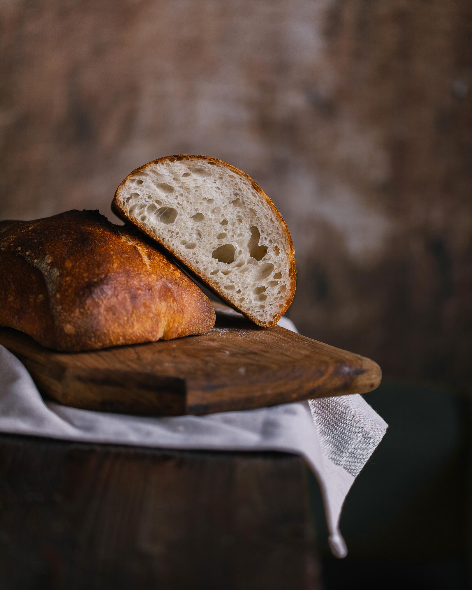 A wooden board with cut bread. A rustic towel lies on a dark wooden table. On it is a wooden board with cut bread. The bread has a crispy crust and beautiful large holes.